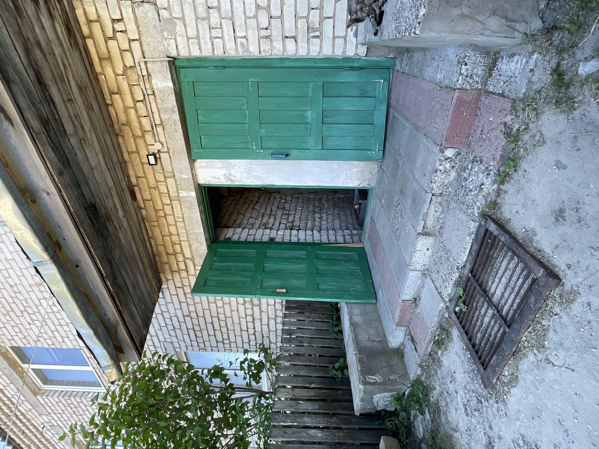 The image shows the entrance to a building with light-colored brick walls. Two green wooden doors are visible. The left door is open, revealing a dark interior. The right door is closed. A wooden overhang with a metal gutter extends over the entrance. A wooden fence and a tree are to the left of the building. The ground in front of the entrance is paved with square tiles, and a metal grate is visible on the ground. The scene appears to be an exterior view of a building in a residential area, possibly in the city of Krasnye Baki, Russia.
