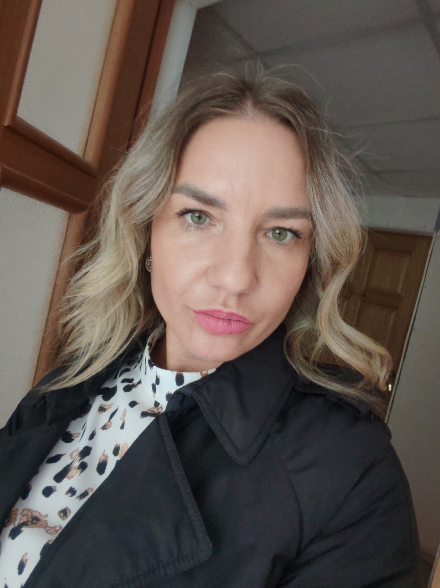 A blonde woman with green eyes and pink lipstick is posing for a selfie. She is wearing a black coat over a white top with a brown and black animal print. The background includes a wooden door frame and a partial view of a wooden door. The location is not identifiable from the image, and it is impossible to determine if it is in Petropavlovsk-Kamchatsky, Russia.