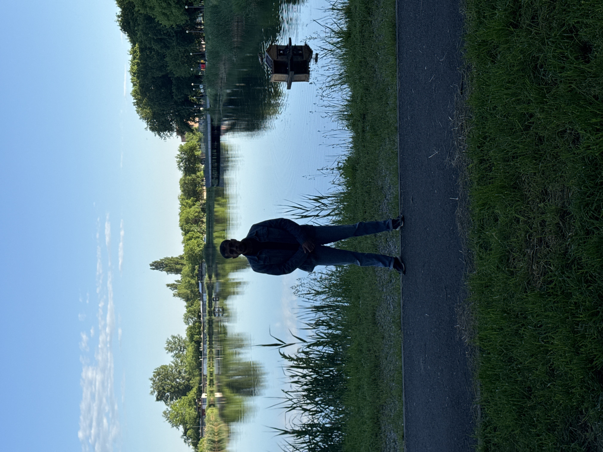 A full body shot of a person standing on a paved path next to a body of water. The person is wearing a dark denim jacket, blue jeans, and dark shoes. Green grass grows on the right side of the path. Reeds grow in the area between the path and the water. A still body of water reflects the sky and trees on the opposite shore. A small wooden structure floats on the water. The image appears to have been taken in Unknown, Unknown.