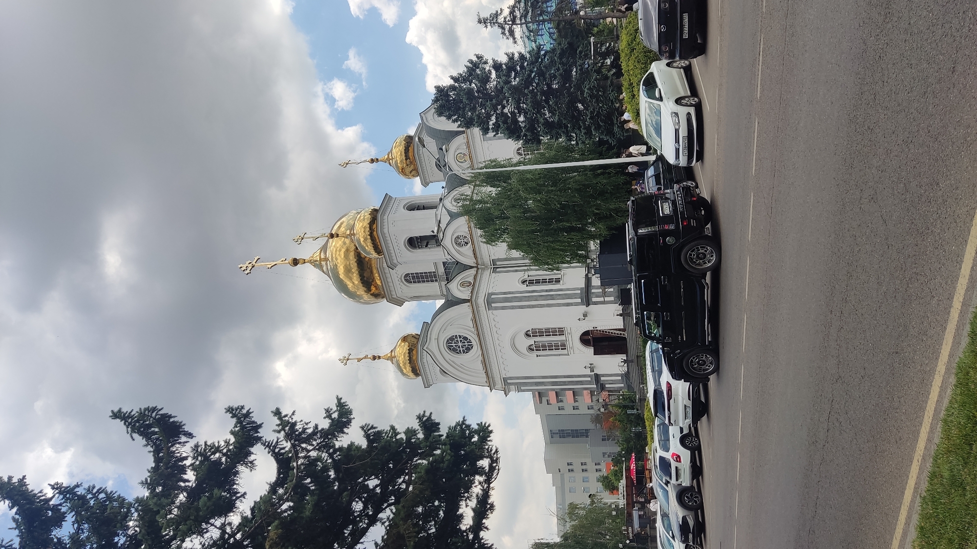 The image displays a side view of a white church with golden domes, topped with crosses. The church has multiple windows and intricate architectural details. Adjacent to the church, parked along the side of a road, are several cars, including a black SUV and a white sedan. Some people are visible near the church and the cars.
The scene appears to be outdoors, with a cloudy sky that suggests either early morning or late afternoon. Trees surround the church, with one tall evergreen tree prominently featured in the foreground. The road is paved and has white lane markings, indicating it is a public street. Some buildings are visible in the background, suggesting an urban or suburban setting. There is some text visible on the license plates of the cars, but it is too blurry to read. The overall impression is of a serene, possibly spiritual location on a somewhat overcast day.