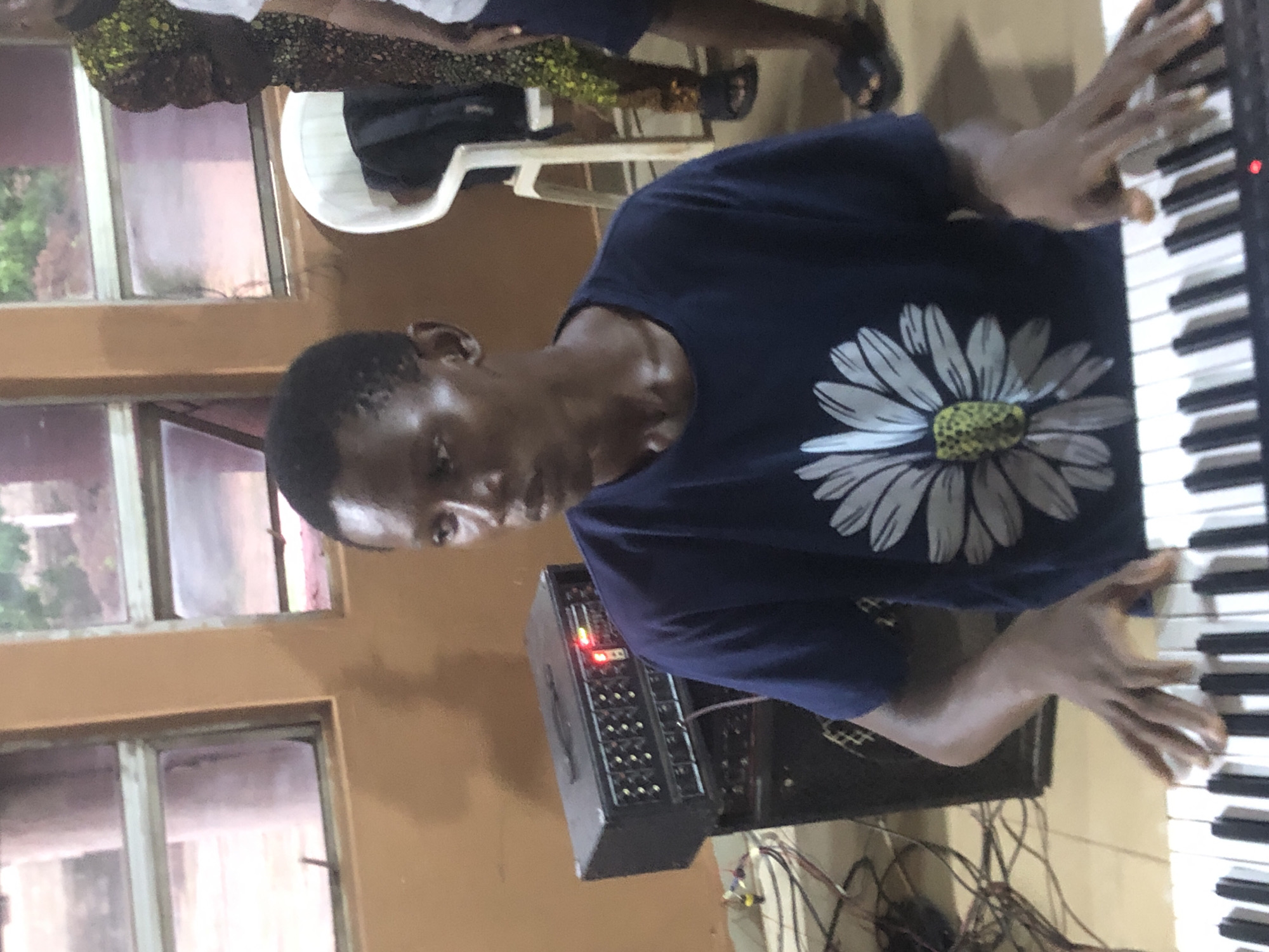 A young Black male with short hair and a dark blue t-shirt featuring a large white and yellow daisy design is seated in front of a keyboard, his hands positioned over the keys, suggesting he is playing. Behind him, a black amplifier with a red light on its control panel is stacked on another speaker. The background shows windows with blinds, an orange wall, and parts of other individuals, one wearing a patterned garment and the other in a white top and dark shorts. The scene appears to be indoors, possibly in a community hall or church in Nsugpe, Nigeria.