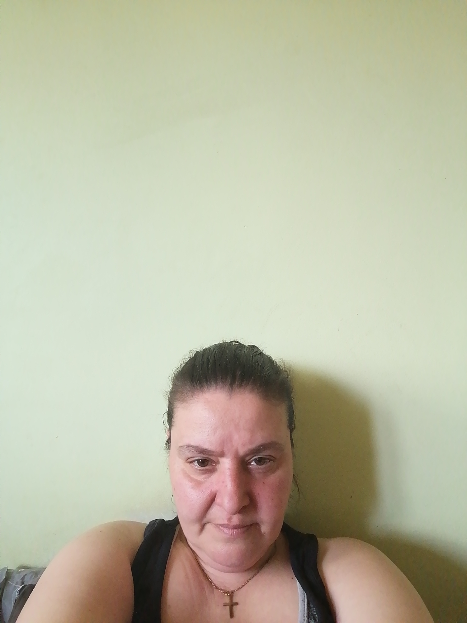 A medium shot depicts a middle-aged woman with fair skin and dark hair pulled back from her face. She is wearing a black tank top and a gold necklace with a cross pendant. The background is a plain, light green wall. The location is unknown.