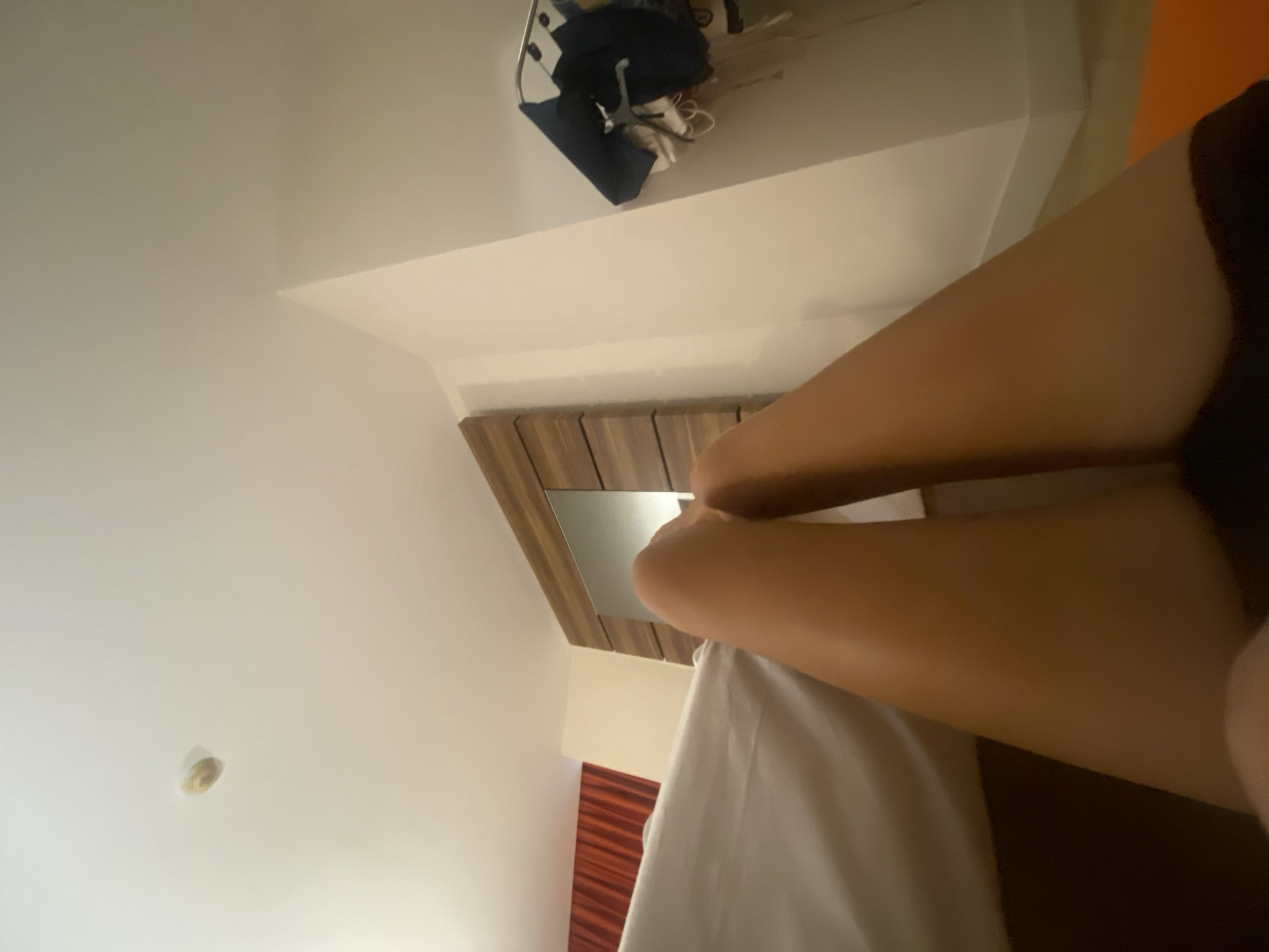 The image depicts a low-angle view of a person's legs lying on a bed in a hotel room. The room features a white ceiling with a circular light fixture and a wooden headboard with a mirror. A red curtain is visible in the background to the left. To the right, a rack of clothes hangs on the wall. The lighting is dim, creating shadows and a warm ambiance. The city of Aksu, Turkey, is not depicted in this image, nor is it relevant to the visual content.