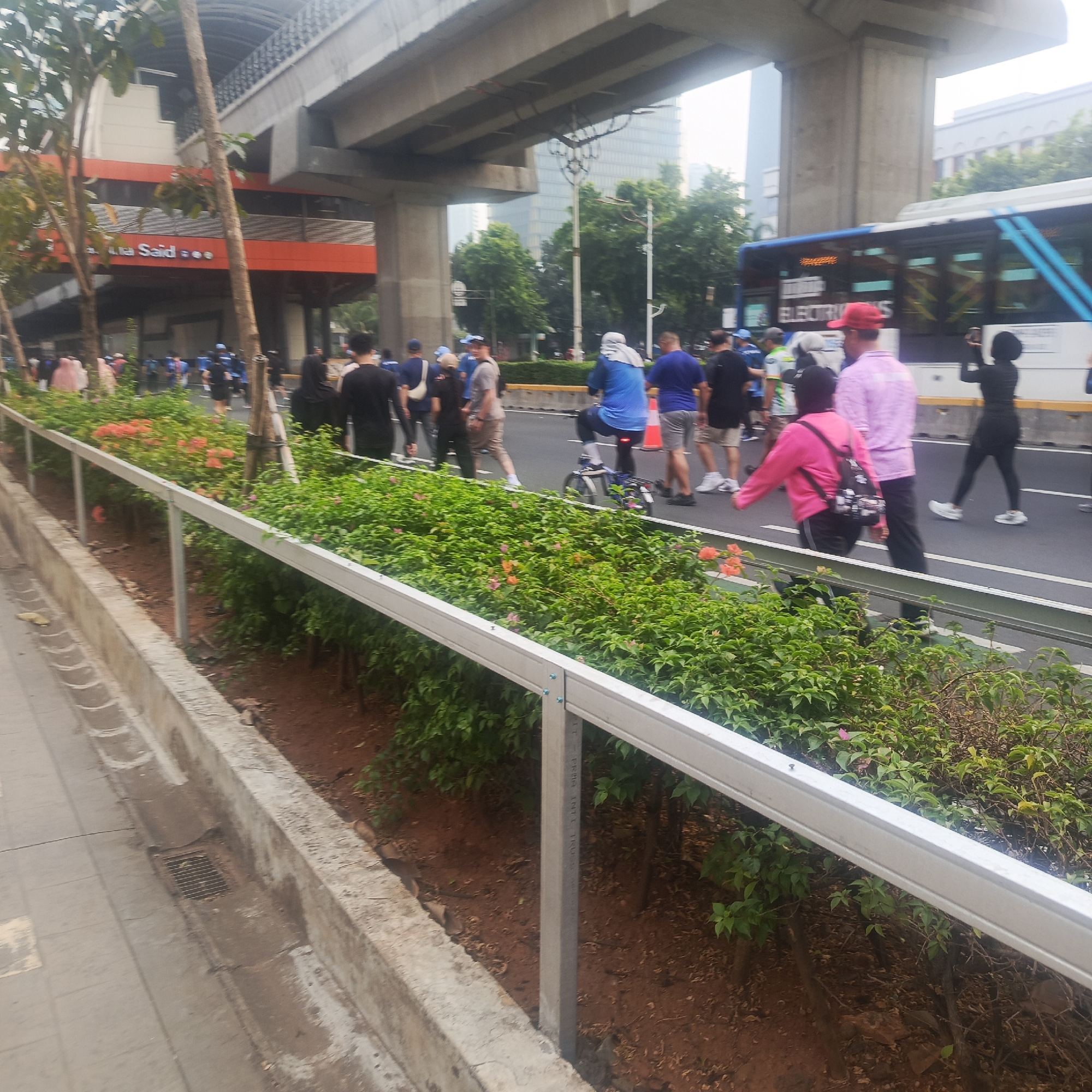 The image captures a street scene in South Jakarta, Indonesia, likely near a transportation hub, given the elevated structure that appears to be a mass transit line. The foreground is dominated by a manicured garden with lush green foliage and scattered pink and orange flowers, bordered by a white metal railing. A concrete walkway is visible to the left, featuring a drainage grate.
In the background, a white bus with blue accents, displaying "ELECTRICITY" on its side, is in motion. Several people are walking on the road, some appearing to be participants in an event or gathering. Among them, a woman in a pink jacket and a backpack is walking towards the viewer. Another woman in a black hijab and outfit is seen taking a photograph with her phone. A person in a wheelchair is also present on the road.
The elevated infrastructure, a common sight in urban areas like South Jakarta, casts shadows over the scene. The overall atmosphere suggests a public space with ongoing activity, possibly related to a community event or daily commuting.