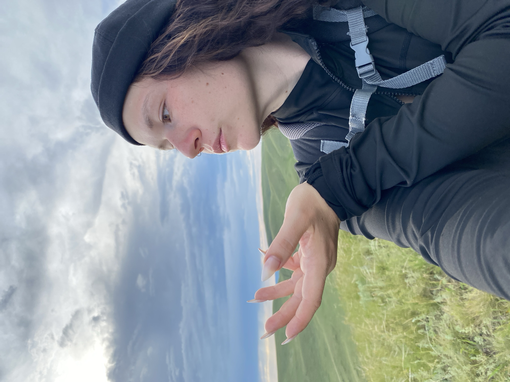 A young woman wearing a black beanie and a black zip-up jacket with a backpack on is looking to the side with her hand outstretched in front of her. She has long, light-colored fingernails. She is outdoors with a landscape of rolling green hills and a cloudy sky in the background. The location is Verkhneozernoe, Russia.