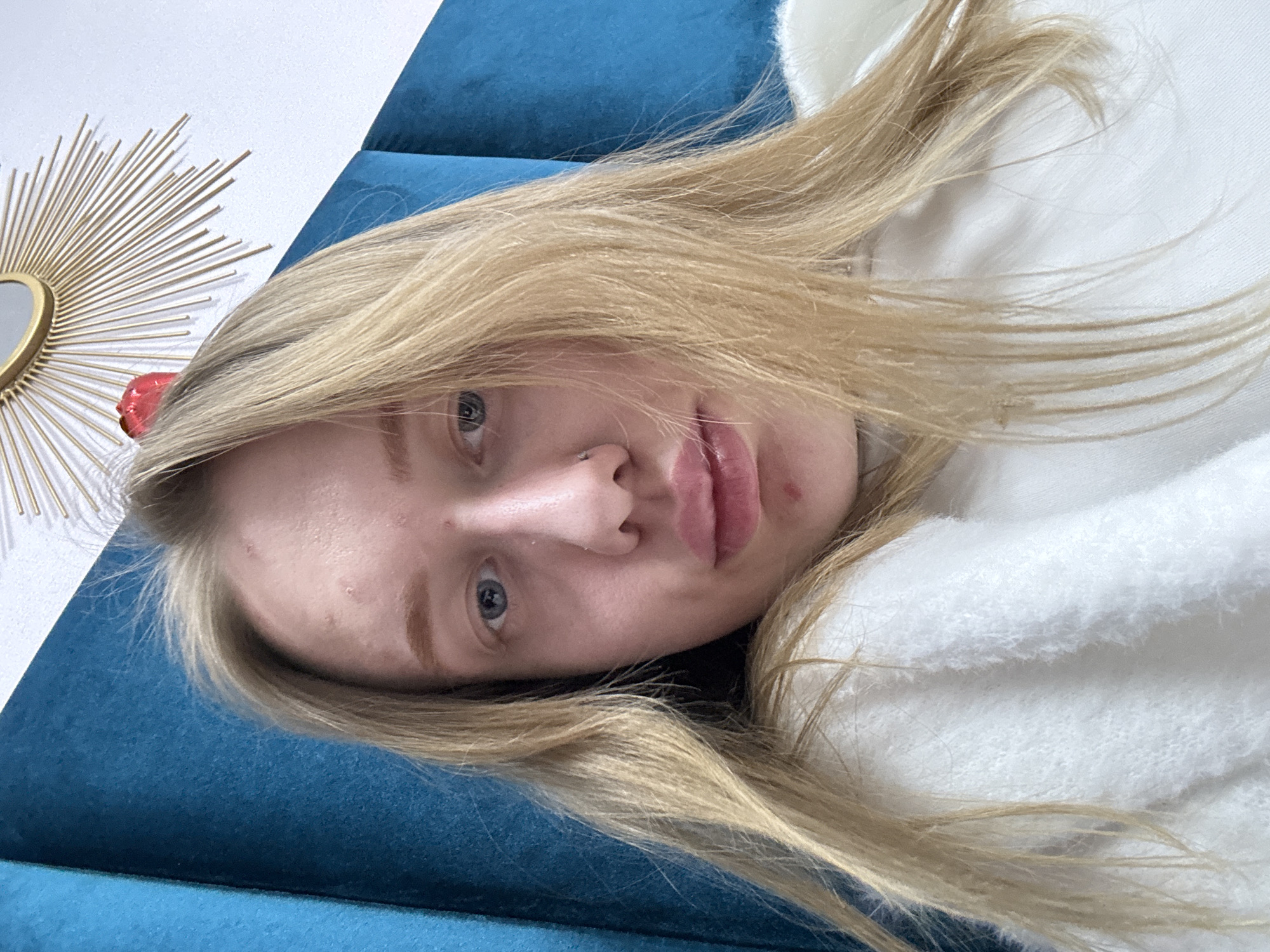 A blonde-haired woman is lying on a blue, textured surface, possibly a couch or bed. She is wearing a white, textured top or sweater. In the background, there's a decorative sunburst mirror attached to a white wall. A red object is visible adjacent to the mirror. There is no verifiable indication of the image being taken in Rostov-on-Don, Russia.