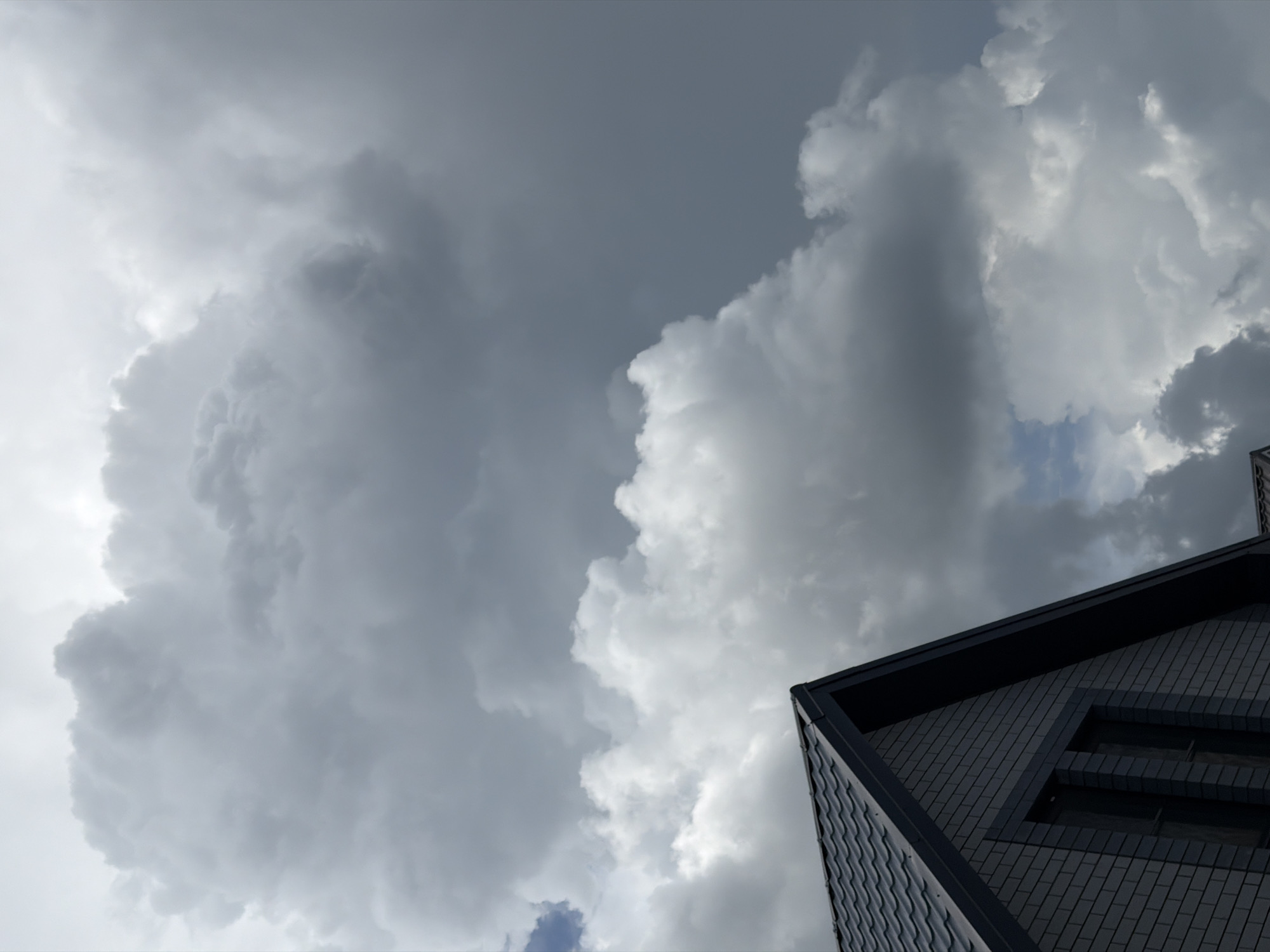 The image shows a low-angle view of a dark-colored building against a backdrop of heavy, grey clouds. The building occupies the lower right portion of the frame, with its corner pointing upwards and towards the left. The building appears to be covered in a patterned surface, possibly tiles or panels. A rectangular window is visible. The sky is dominated by large, billowing clouds. The location is Rossiyskiy, Russia.