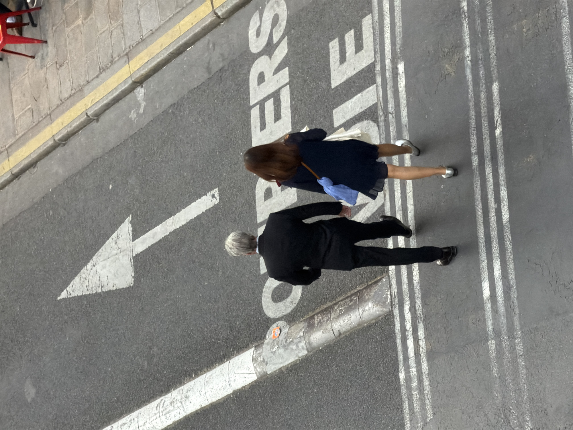 This overhead shot captures two individuals walking on a street in Paris, France. The scene is dominated by a dark asphalt road surface featuring prominent white road markings, bordered by a light-colored paved sidewalk to the left.
A woman with long brown hair, dressed in a dark blue or black dress or tunic, is seen walking with silver low-heeled shoes. She holds a light blue item and a white item, possibly bags or folders, in her hands. Ahead of her is a man with grey hair, wearing a black suit and dark shoes. Both individuals appear to be walking in the same general direction, broadly aligned with a large white arrow painted on the road, which points towards the left of the frame.
The road features several distinct white markings: the large directional arrow, partially visible text that reads "COUPERS DE PIE" (likely part of a French instruction such as "COUPERS DE PIED," meaning "cross on foot," indicating a pedestrian area), and multiple parallel white lines extending across the road, possibly delineating a specific lane or crossing. A grey cylindrical bollard with a small orange sticker is visible near the center of the road. To the far left, a section of a paved sidewalk with a yellow curb line is visible, along with the red legs of a chair, suggesting an adjacent cafe or outdoor seating area.
The image appears to be taken during the daytime, under clear weather conditions, indicated by the bright, even lighting and the absence of shadows that would suggest harsh midday sun or late evening. The French text on the road serves as a clear location cue for Paris.