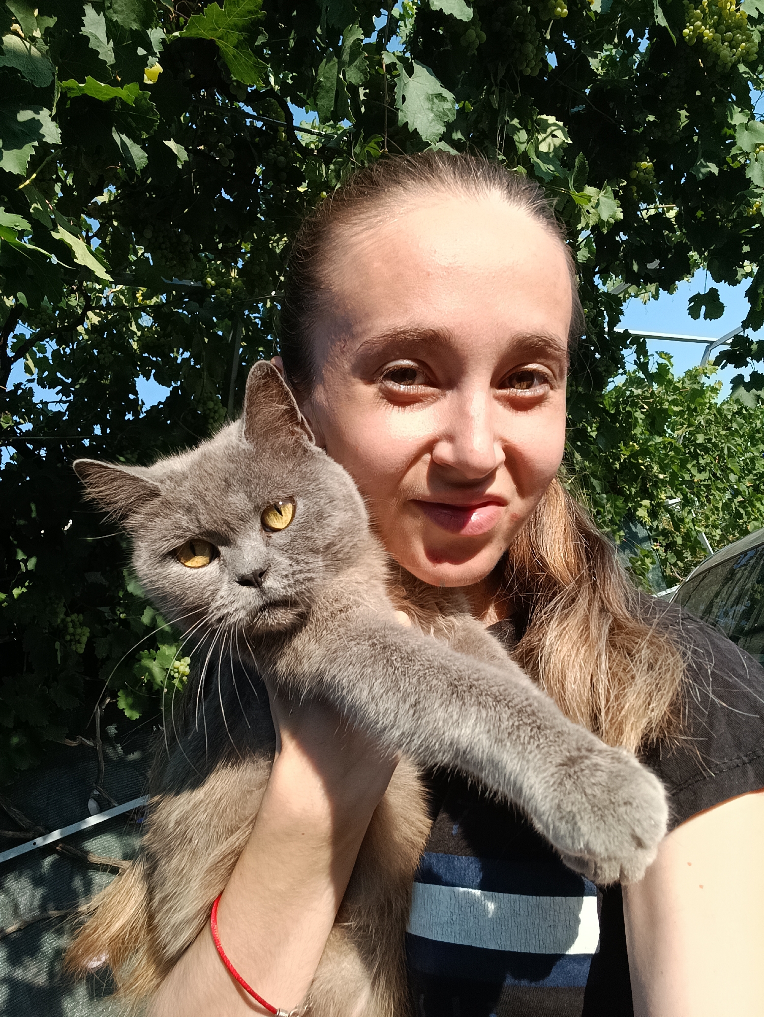 A woman with light brown hair pulled back is holding a gray cat in front of a green leafy vine and a bright blue sky. The woman is looking towards the camera and smiling slightly. The cat, with yellow eyes, is looking towards the left of the frame. The woman is wearing a dark shirt with a blue and white striped pattern visible. The scene appears to be outdoors in a rural setting, possibly a garden or backyard, given the presence of the vine and the overall natural lighting. The image was taken in Vypasne, Ukraine.