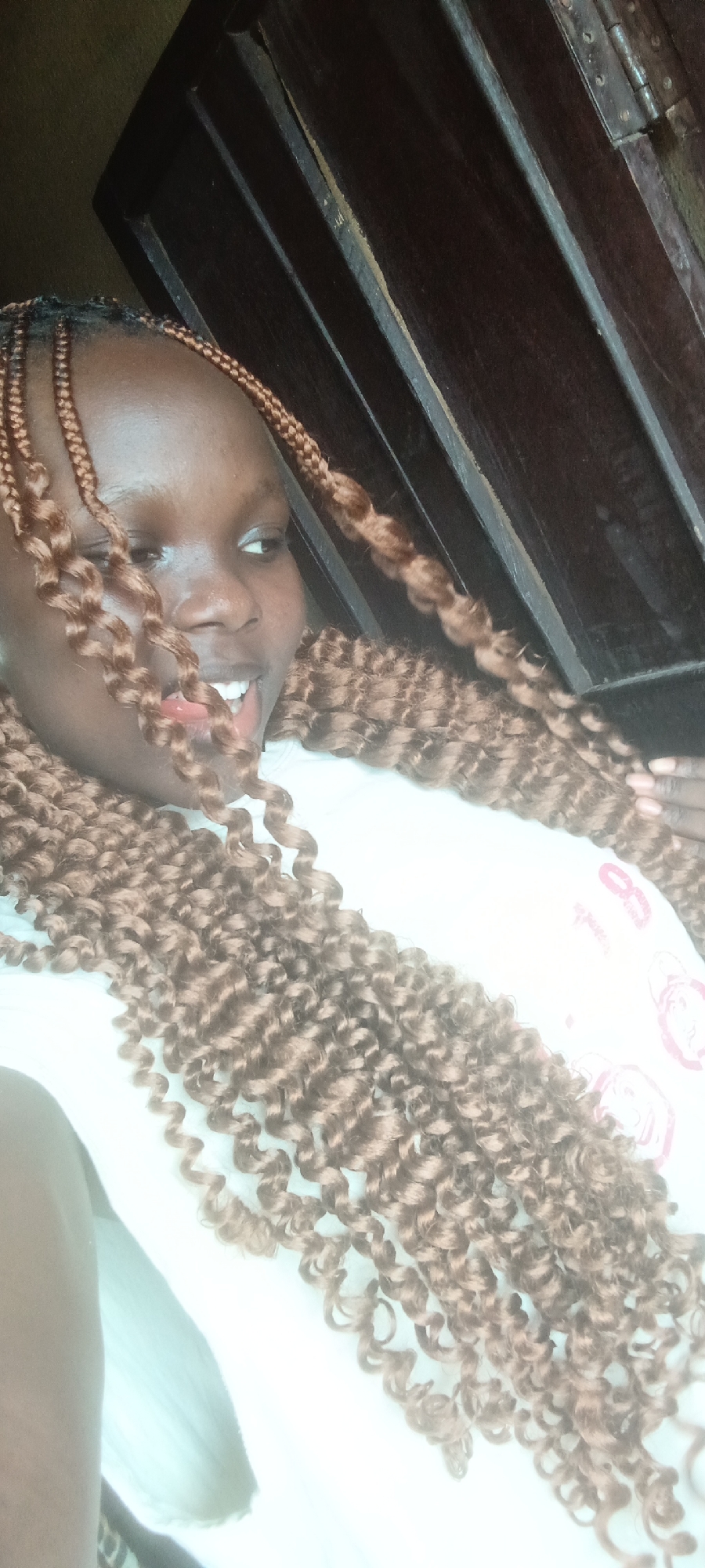 A vibrant portrait captures a young woman, likely in Onitsha, Nigeria, against a dark wooden background. She has dark skin and is adorned with long, voluminous, light brown, curly braided hair, which cascades over her shoulders and chest. The braids feature tightly coiled curls, creating a striking texture. She is wearing a simple white t-shirt, on which a faint, faded red graphic or text is partially visible on her right side.
The woman is looking to her right (viewer's left) with a wide, open-mouthed smile, revealing her teeth and a glimpse of her tongue. Her animated expression conveys joy, laughter, and a relaxed, playful mood. She appears to be in an indoor setting, possibly reclining or lying down, given the angle of the shot. The background consists of dark brown wooden panels, possibly part of a door or furniture, with visible vertical lines and some metal hardware, like a hinge, adding to the interior setting detail. The image has a cheerful and informal ambiance.
