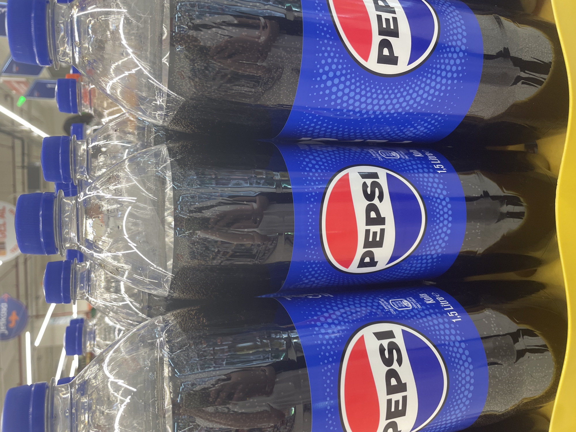 The image shows a close-up, slightly angled view of several plastic bottles of Pepsi soda. The bottles are arranged in a stack, with the labels clearly visible. The Pepsi logo, featuring the iconic red, white, and blue circle with the word "PEPSI" in white lettering, is prominent on each label. The blue portion of the label is decorated with a pattern of small white dots. Text on the labels indicates "1.5 Litre" and "Kola" (Turkish for cola). Some bottles also show nutritional information.
The setting appears to be a retail environment, possibly a supermarket or convenience store, given the presence of lighting fixtures visible in the background and what looks like a store sign with the word "Jeskosa" in the upper left corner. The bottles are likely on a shelf or in a display. The lighting suggests it is daytime, as might be expected in a store. There are no people visible in the image, and the focus is entirely on the product. The overall impression is one of a commercial display of beverages.