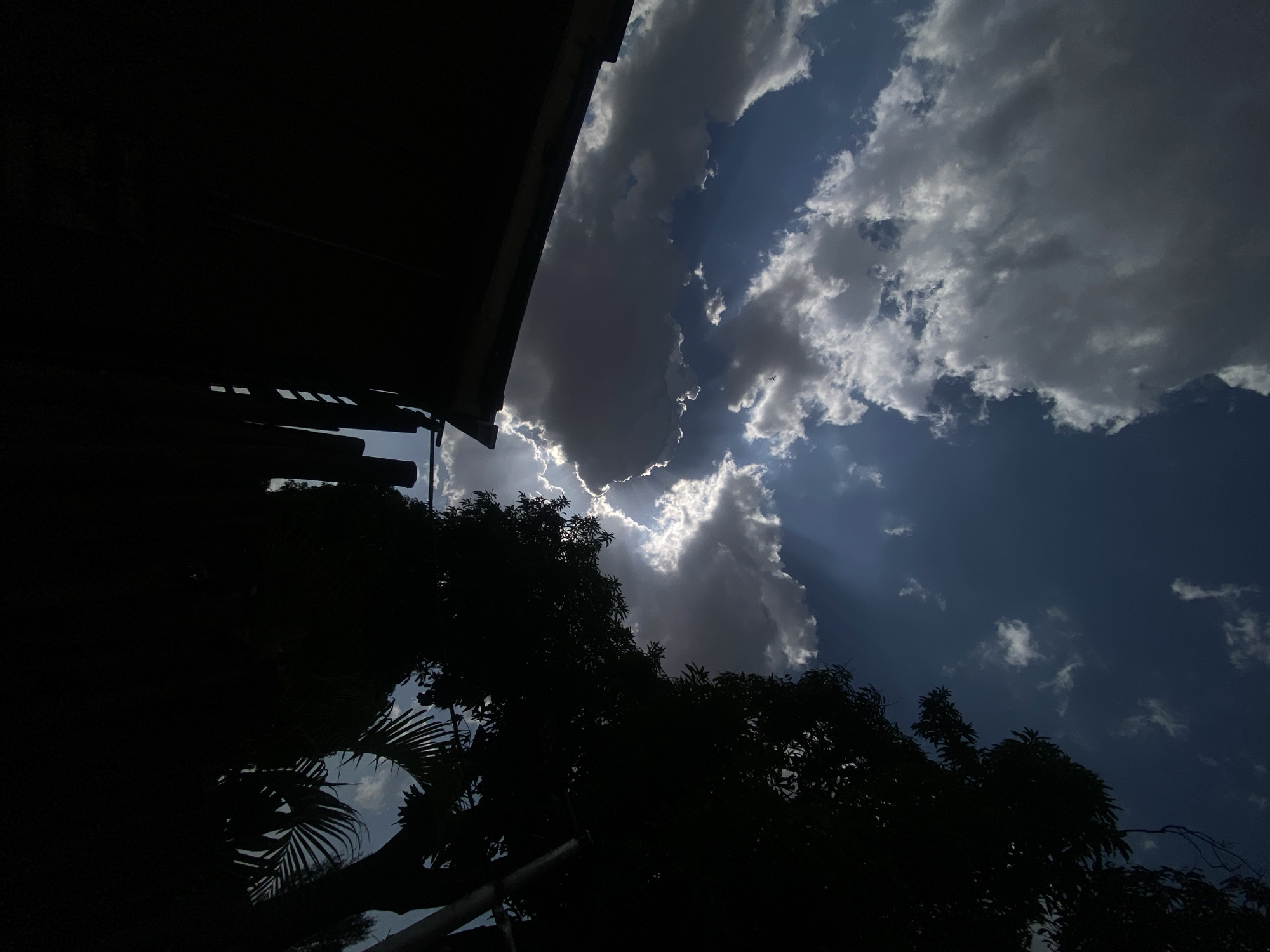 The image is a low-angle shot taken in what appears to be an outdoor setting, possibly in Barden Yana, Nigeria. The composition is divided between dark silhouettes of trees and a building structure in the foreground and a dynamic sky filled with clouds in the background. The sky features bright patches where sunlight breaks through the clouds, creating a dramatic contrast between light and shadow. The clouds vary in density, with some appearing as dark, heavy formations and others as lighter, scattered wisps. The building structure on the left-hand side is mostly obscured by shadow, but hints of its construction, possibly including a roof and window or vent-like openings, are visible. The trees are represented as dark silhouettes, their leaves and branches obscuring much of the lower portion of the image. The scene lacks the presence of people or explicit actions, focusing primarily on the interplay of natural elements and architectural forms.