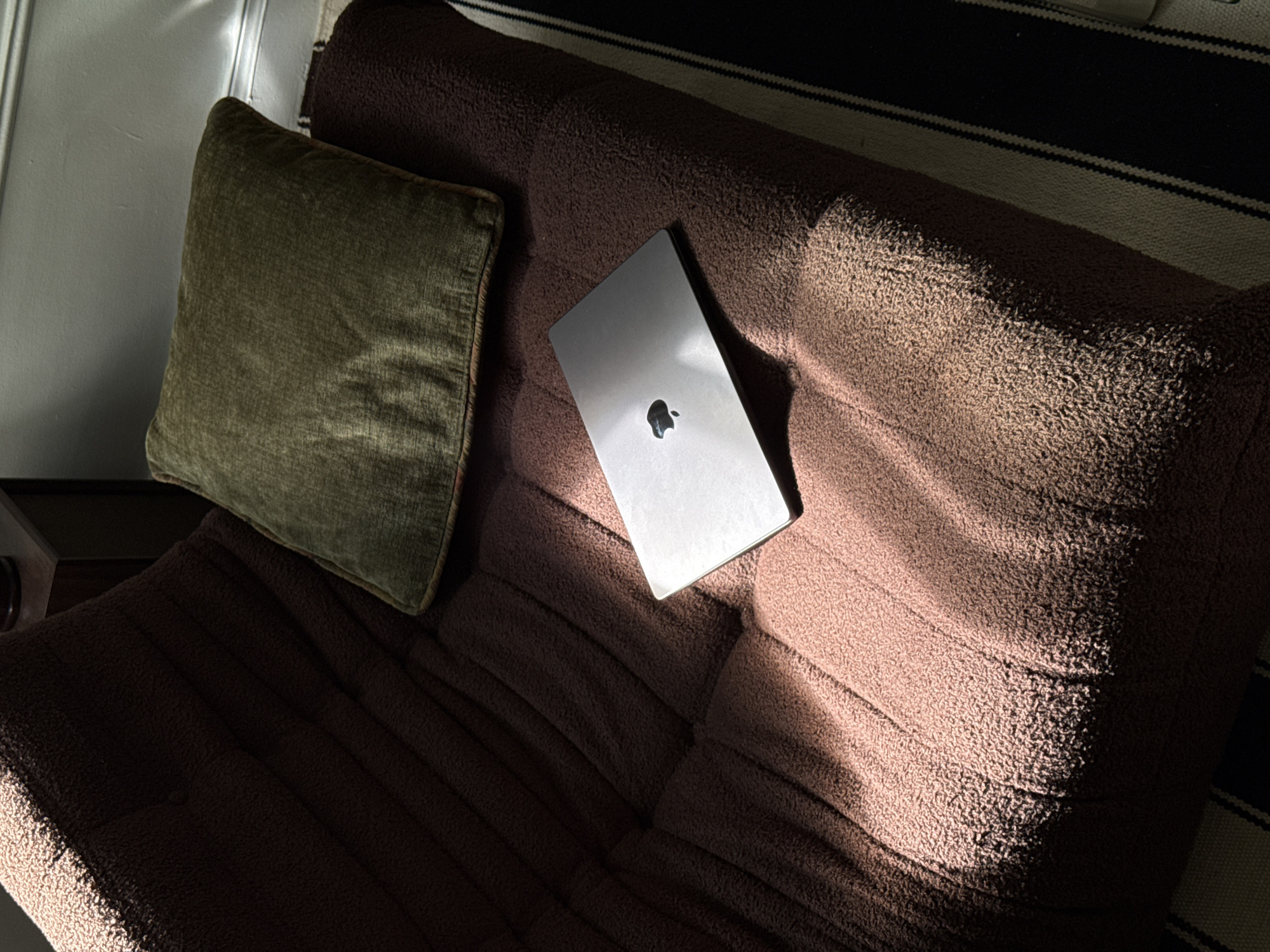 The image shows a laptop resting on a brown, textured couch with a green pillow to the left. The couch appears to be a futon, with visible ridges and texture. Sunlight casts shadows across the scene. The background includes a striped rug and part of a wall. There is no evidence to confirm that this image was taken in the city of Montreal, Canada.