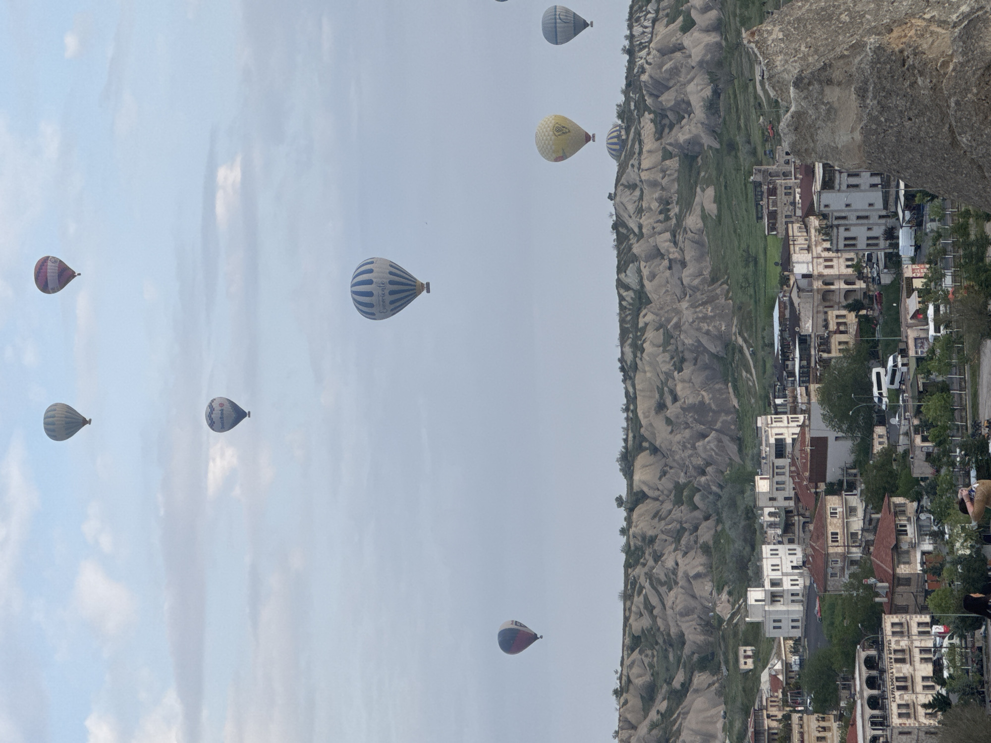 The image captures a scene in Göreme, Turkey, known for its unique landscape and hot air balloons. The sky is filled with hot air balloons of various colors and patterns, some with visible branding. Below, the terrain consists of distinctive rock formations and cave dwellings, typical of Cappadocia. Houses and buildings are nestled among the rocks, indicating a settlement area. The photograph is taken from an elevated position, providing a wide view of the valley.