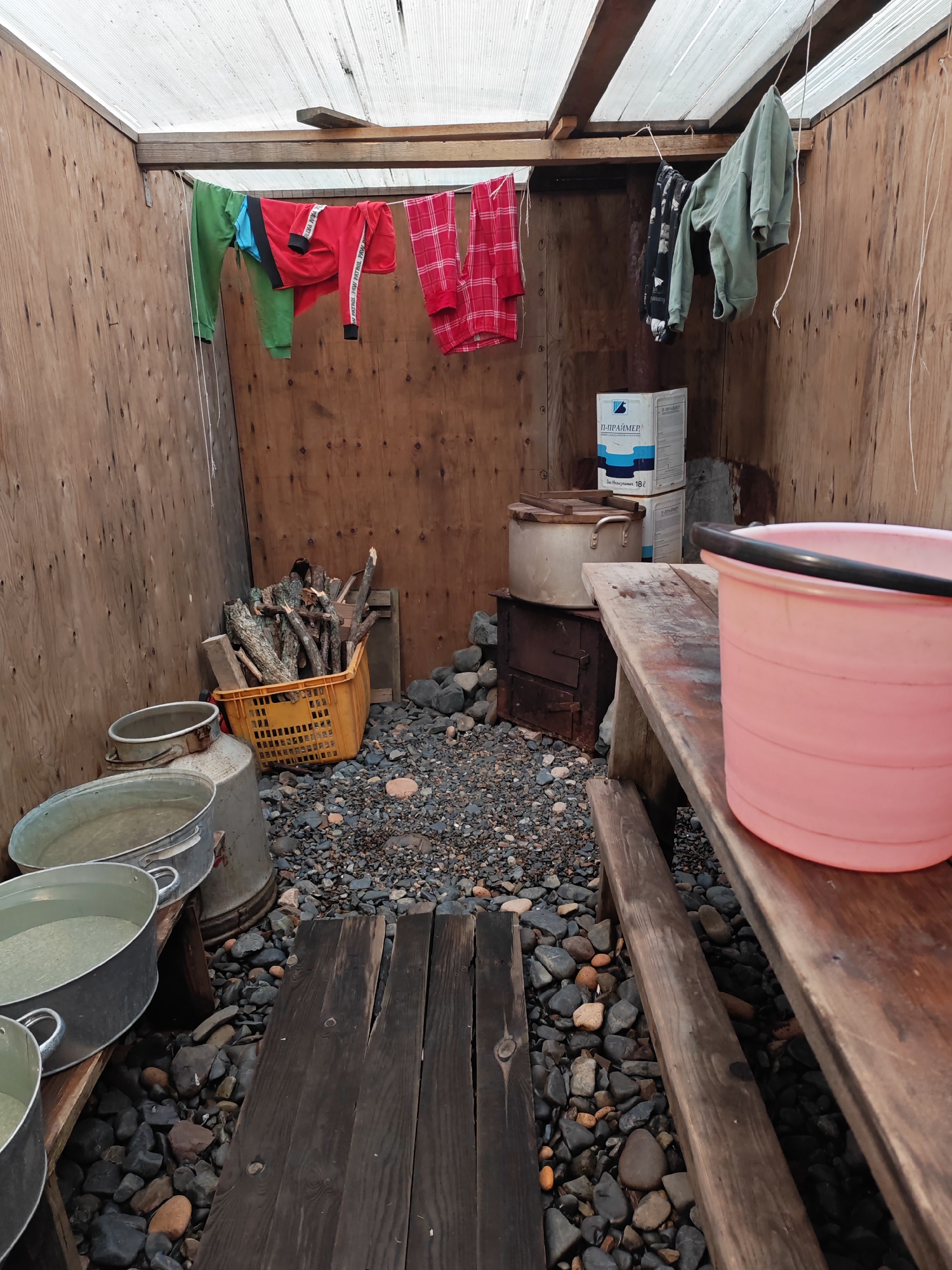 This image depicts an outdoor storage area in Reineke, Russia, constructed with wooden walls and a translucent plastic roof. In the center of the scene, a collection of firewood is stored in an orange plastic basket, next to a small metal wood-burning stove. Various metal tubs, some containing water, are arranged on the left side of the space. Clothes are drying on a line suspended from the ceiling, adding a domestic touch to the utilitarian setting. A wooden bench runs along the right side of the image, topped with a large pink bucket. The ground is covered with gravel and stones, typical of outdoor areas. The overall impression is one of a practical, possibly temporary, shelter used for storage and possibly basic living needs.