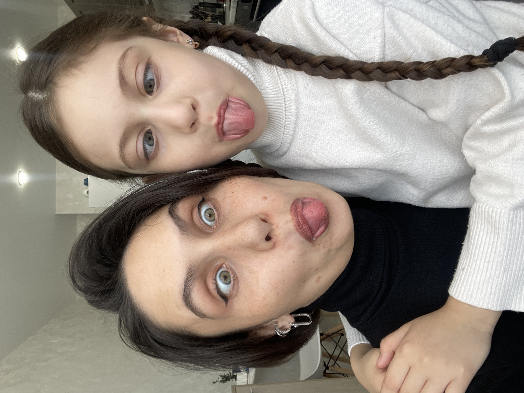 Two females are posing for a selfie. The older woman has short dark hair and is wearing a black turtleneck sweater. She has green eyes and is sticking her tongue out. The younger girl, likely a child, is above her, wearing a white turtleneck sweater and has a brown braid. She is also sticking her tongue out. They are likely indoors in Izhevsk, Russia.