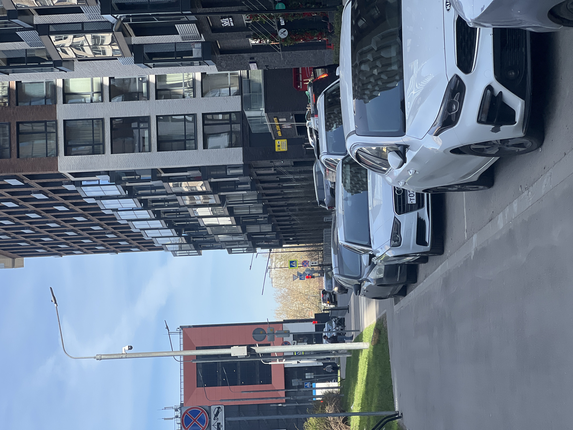 An urban street scene unfolds on a clear, sunny day, featuring modern architecture, parked vehicles, and street infrastructure. The image appears to be taken from street level, looking down a thoroughfare.
On the left, a series of multi-story, contemporary buildings with intricate facades dominate the background. These structures feature numerous windows, some reflecting the sky and surrounding buildings, and various textural patterns in their design, suggesting residential or office use.
To the right, a row of cars is parked parallel to the street. The most prominent vehicle in the foreground is a white Kia Stinger. Behind it, parts of other white and darker-colored cars are visible, including one with an Audi logo. The cars are parked on a paved surface next to a narrow strip of green grass.
Further down the street, beyond the parked cars, the road continues, with other vehicles partially visible in the distance. A lower-rise building with a reddish-orange facade and large dark windows stands on the right side of the street, contrasting with the taller buildings opposite.
Street furniture includes a tall lamppost with a security camera attached, and various traffic signs. A "no parking" sign (red circle with diagonal line over a 'P') is visible in the lower right. Other signs indicate a pedestrian crossing (yellow triangle with walking person) and directional information. Text visible on building signs includes "Shell Beauty Salon" (partially obscured) and a yellow sign with "ПАРКОВКА" (Parking) and "Аренда" (Rent). Another sign features a spoon and fork icon, indicating a restaurant. The blue sky above suggests good weather conditions.