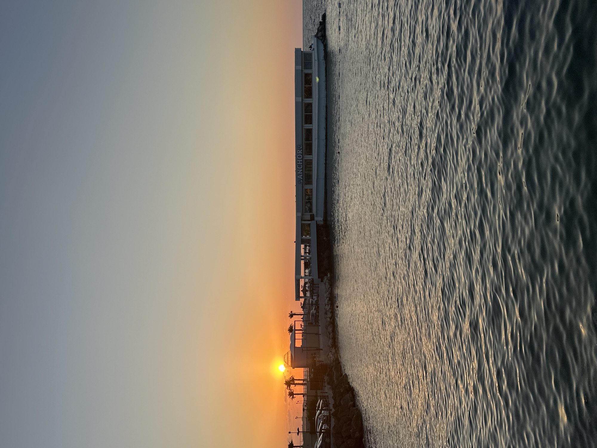 The image depicts a tranquil scene at sunset in Kyrenia, Northern Cyprus. The sun, a bright orange orb, is low on the horizon, casting a warm glow over the sky and reflecting on the calm sea. In the foreground, the dark blue water is gently rippled, with sunlight glinting off its surface.
Along the coastline, a rocky breakwater leads to a modern-looking building complex situated over the water. This complex appears to be a restaurant or bar, with large windows overlooking the sea and the name "ANCHOR" clearly visible on its facade. Several cars are parked in a lot adjacent to the water's edge, suggesting a public area or a place of business. Palm trees dot the landscape, adding a tropical feel to the setting. A few people are visible near the building and on the breakwater, enjoying the evening ambiance. The sky transitions from a pale orange near the horizon to a soft blue at the top of the frame, indicating the fading light of dusk.