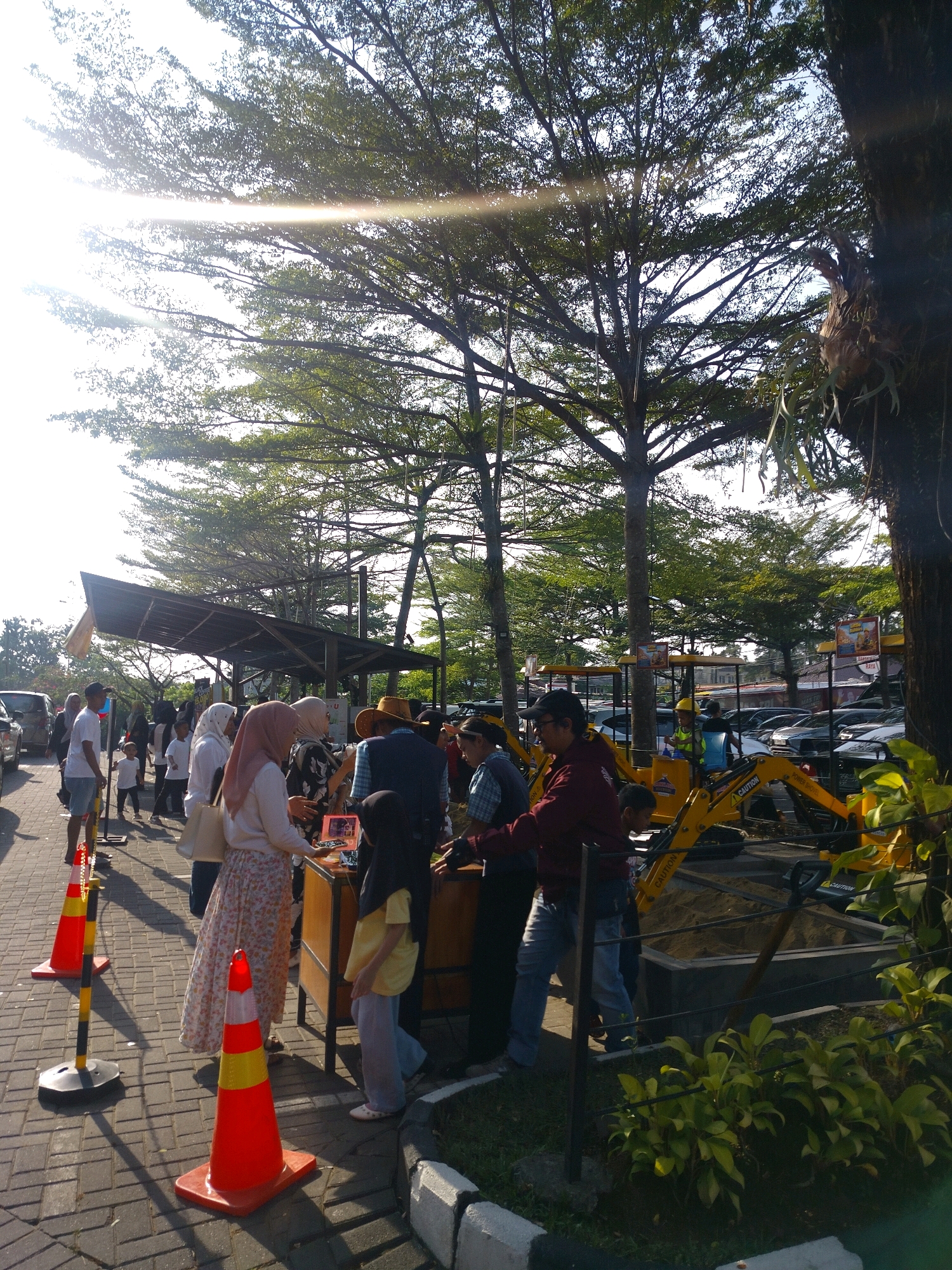 In a sunlit outdoor setting in Megamendung, Indonesia, a group of people gather near a temporary stall or display. The background features lush green trees with sunlight filtering through their branches, creating a dappled effect. Several cars are parked behind the gathering, indicating a public area or event. In the foreground, orange traffic cones mark a boundary or pathway.
People of various ages are present, some dressed in casual attire, while others wear hijabs. A woman in a pink hijab and a floral skirt stands near a table with a laptop, seemingly engaged in a transaction or discussion. A young girl in a yellow shirt and light blue pants stands beside her. Another woman in a white hijab and a white top is also nearby. Further back, a man in a white t-shirt and shorts stands next to a child. A man wearing a maroon jacket and jeans is seen in the mid-ground, bent over what appears to be a construction-themed exhibit with a yellow excavator. Another person, possibly a child wearing a yellow vest and hard hat, is visible behind the excavator. The ground is paved with brick-like stones, and there is a patch of green vegetation to the right of the scene. A large tree trunk is prominent on the far right, with some hanging vegetation and a weathered wooden object attached to it.