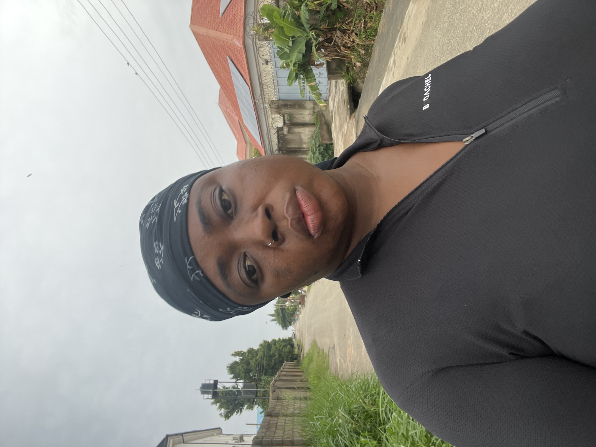 A close-up selfie shows a dark-skinned person with a bandana, likely a woman, wearing a black top with a zipper. The text "B. DACHEL" is visible on the upper part of the top. The person has a nose ring. Behind the person is a residential street in Abuja, Nigeria, indicated by the presence of buildings with corrugated metal roofs and concrete walls. The weather appears overcast. The road surface is light-colored and shows signs of wear. Green vegetation and low brick walls line the street. Electrical wires span across the sky above.