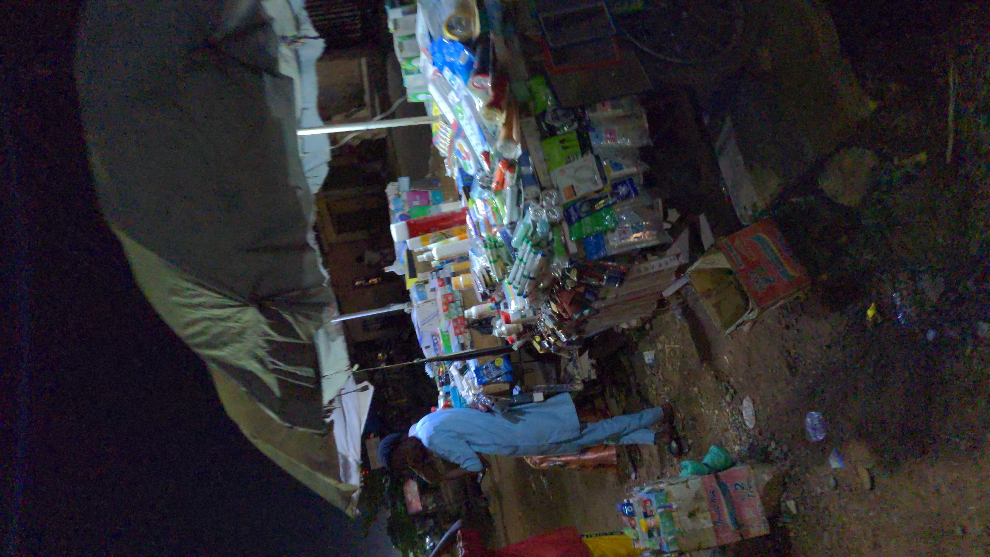 A nighttime scene in Ado, Nigeria, features a man in a light blue outfit standing near a makeshift stall or display of merchandise. The stall contains numerous small items, possibly toiletries or packaged goods, neatly arranged. The area around the stall appears to be somewhat unkempt, with debris and cardboard boxes visible on the ground. A large canvas or tarp-like structure looms to the left, possibly providing shelter or acting as a backdrop for the stall. The environment is dimly lit. The man appears to be interacting with something at the stall.