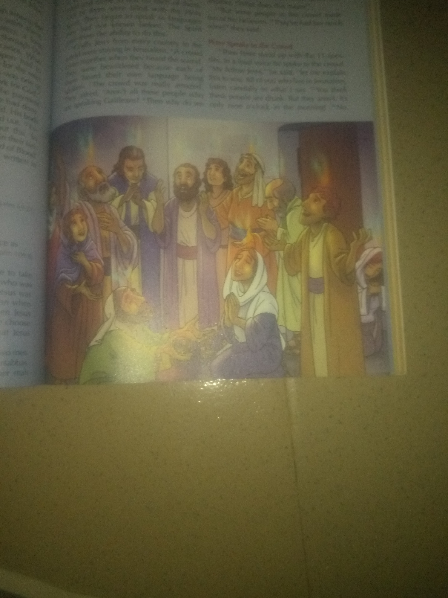 The image is a photograph of an open book with text and an illustration. The illustration depicts a group of people, appearing to be in ancient times, with some figures speaking or gesturing. They are dressed in robes and head coverings. The background of the illustration is abstract and colored with tones of yellow and orange, suggesting a spiritual or dramatic event. The text on the left side of the page is partially visible and appears to be from a religious or historical context. The image is taken from a slightly angled, overhead perspective, and the lighting is uneven, with some areas appearing brighter than others, possibly due to a flash. The overall scene is contained within the pages of a book, which is lying on a textured surface. There is no indication or information present in the image that relates to Jauro Yinu, Nigeria.