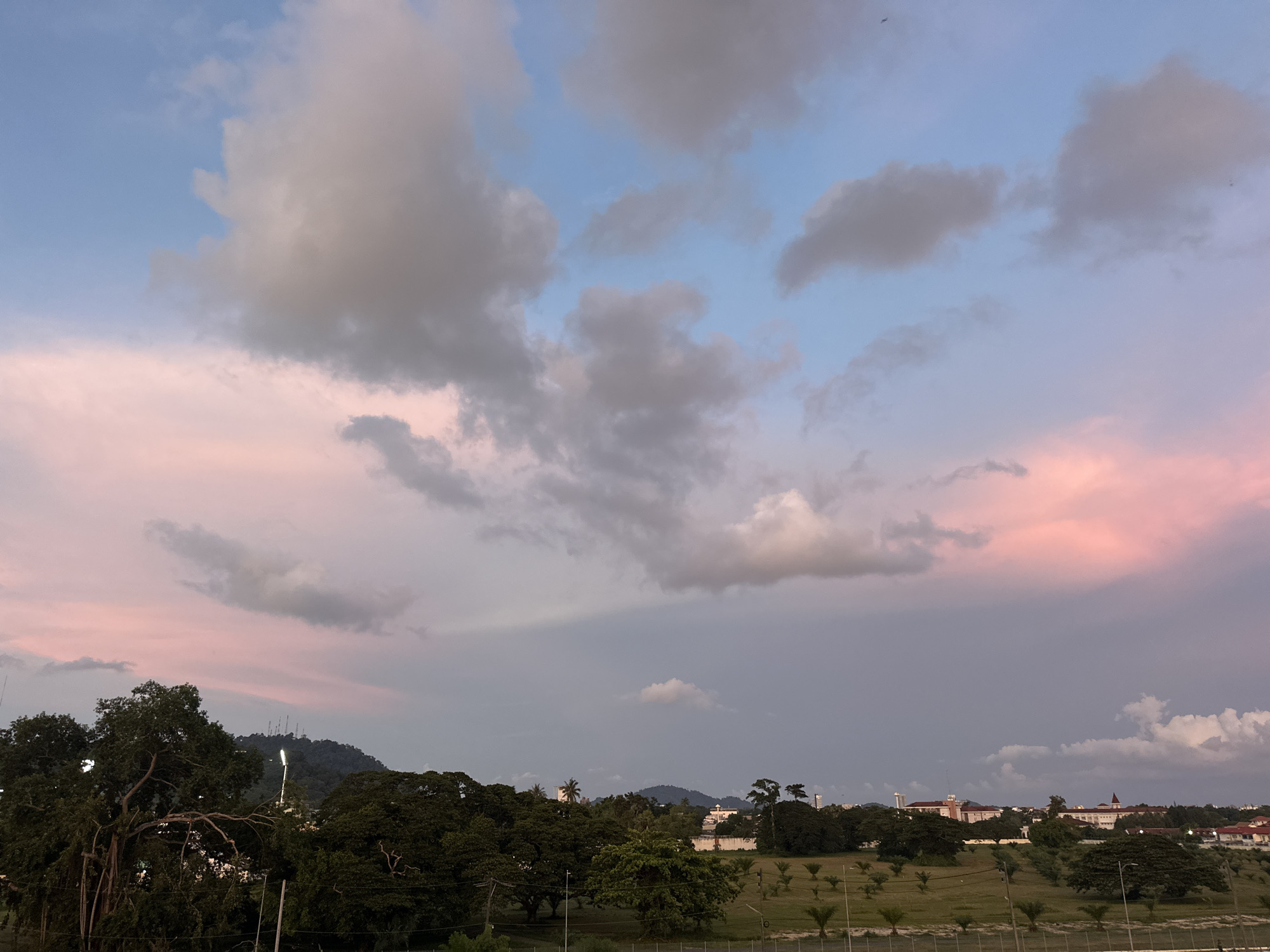 The image depicts a twilight sky over a landscape that includes trees, buildings, and hills. The sky is a mixture of blue, pink, and gray clouds, suggesting it is either sunrise or sunset. In the foreground, there are lush green trees, some appearing to be very large with sprawling branches. Beyond the trees, a line of buildings with red-tiled roofs is visible, situated on a gentle slope. In the distance, hazy hills rise, and the peak of one hill is topped with several communication towers. The lower part of the image shows a grassy field with scattered palm trees, indicative of a tropical or subtropical environment. The overall scene conveys a sense of natural beauty and urban development coexisting. There are no people or animals visible in the image. The location is identified as Wichit, Thailand.