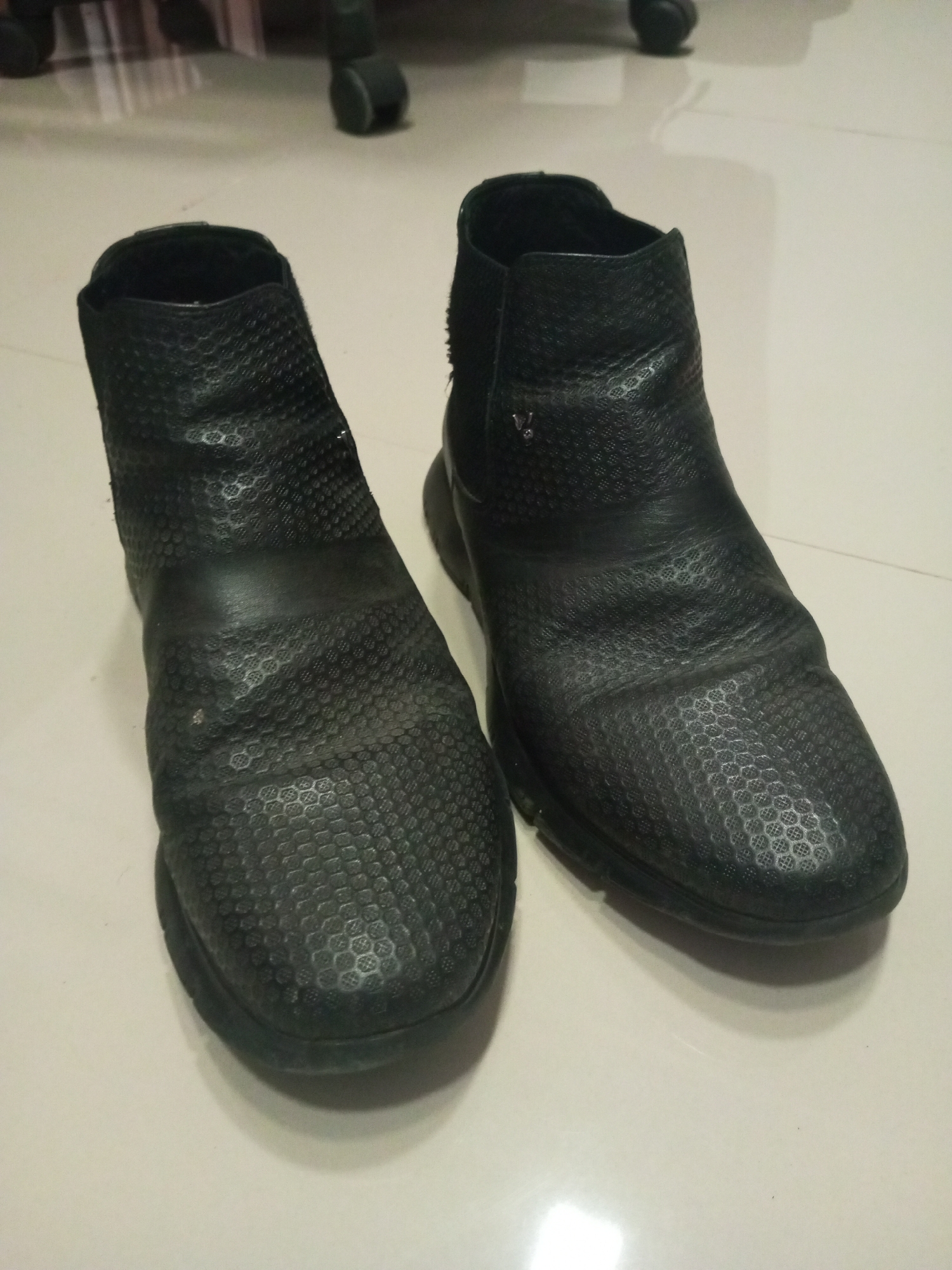 The image shows a pair of used black ankle boots placed on a light-colored tiled floor. The boots are of a similar style, with a textured, possibly leather or synthetic leather, upper featuring a pattern of small circles or hexagons. One boot has a visible metallic emblem or logo, possibly a letter "V," on the side. The boots are wrinkled in places suggesting wear. The environment appears to be indoors, possibly a residence, and the lighting is moderate. There is a blurred object in the background, likely a chair. The image suggests a domestic setting. As there are no indications the photo was taken in Lhokseumawe, Indonesia, the location cannot be determined.