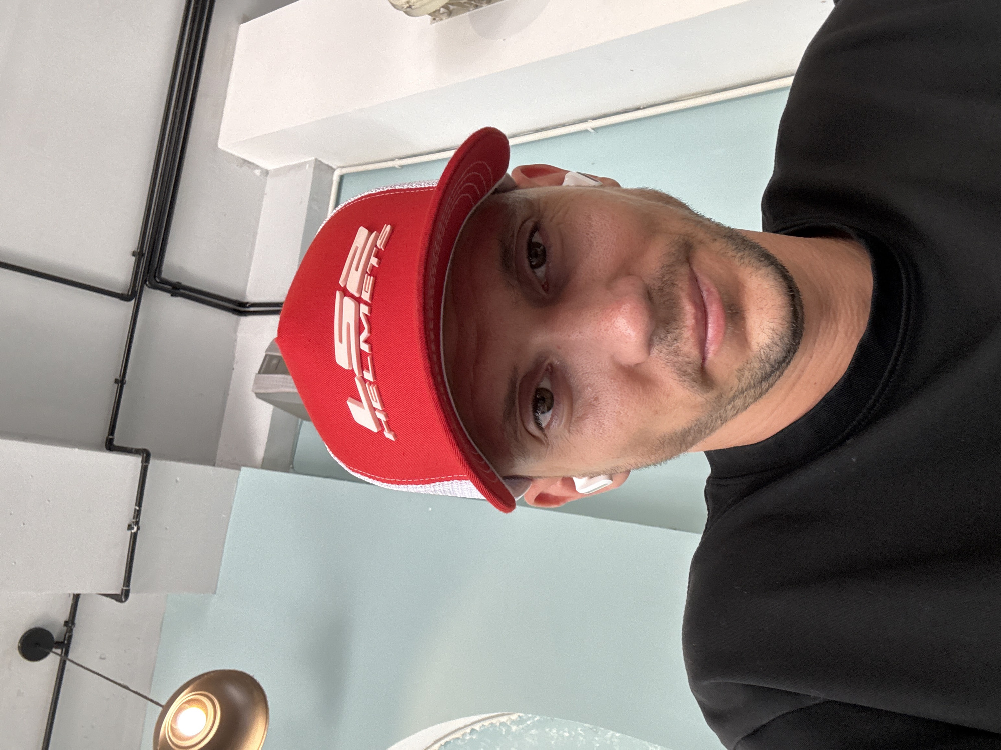 A medium shot shows a person wearing a red cap with a white logo and a black long-sleeved shirt. The person is facing the camera with a slight smile. The background includes a light blue wall, a ceiling with black pipes, and a blurred light fixture. While the location of the city of Nha Trang, Vietnam is not visually indicated in the image, I can not provide that information based on the image alone.