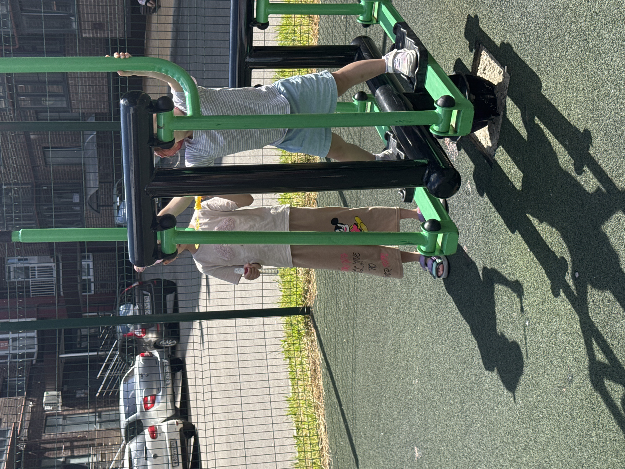 The image shows two children on a playground, likely in Vjazniki, Russia. One child is wearing a striped t-shirt and light blue shorts, with white sneakers. They are positioned on a green and black exercise machine, with their legs extended and hands gripping a green bar. The second child is wearing a light-colored t-shirt and beige pants with Mickey Mouse graphics. They are also on the exercise machine, in a lower position, with their hands on the black bars. The playground surface is a dark green, rubberized material. In the background, there is a chain-link fence, brick buildings, and parked cars, suggesting an urban or semi-urban setting. The presence of shadows indicates it is daytime, likely sunny. The overall mood seems to be one of outdoor recreation. Some text is visible on the second child's pants, including "LOVE TO" and "MR. RA".