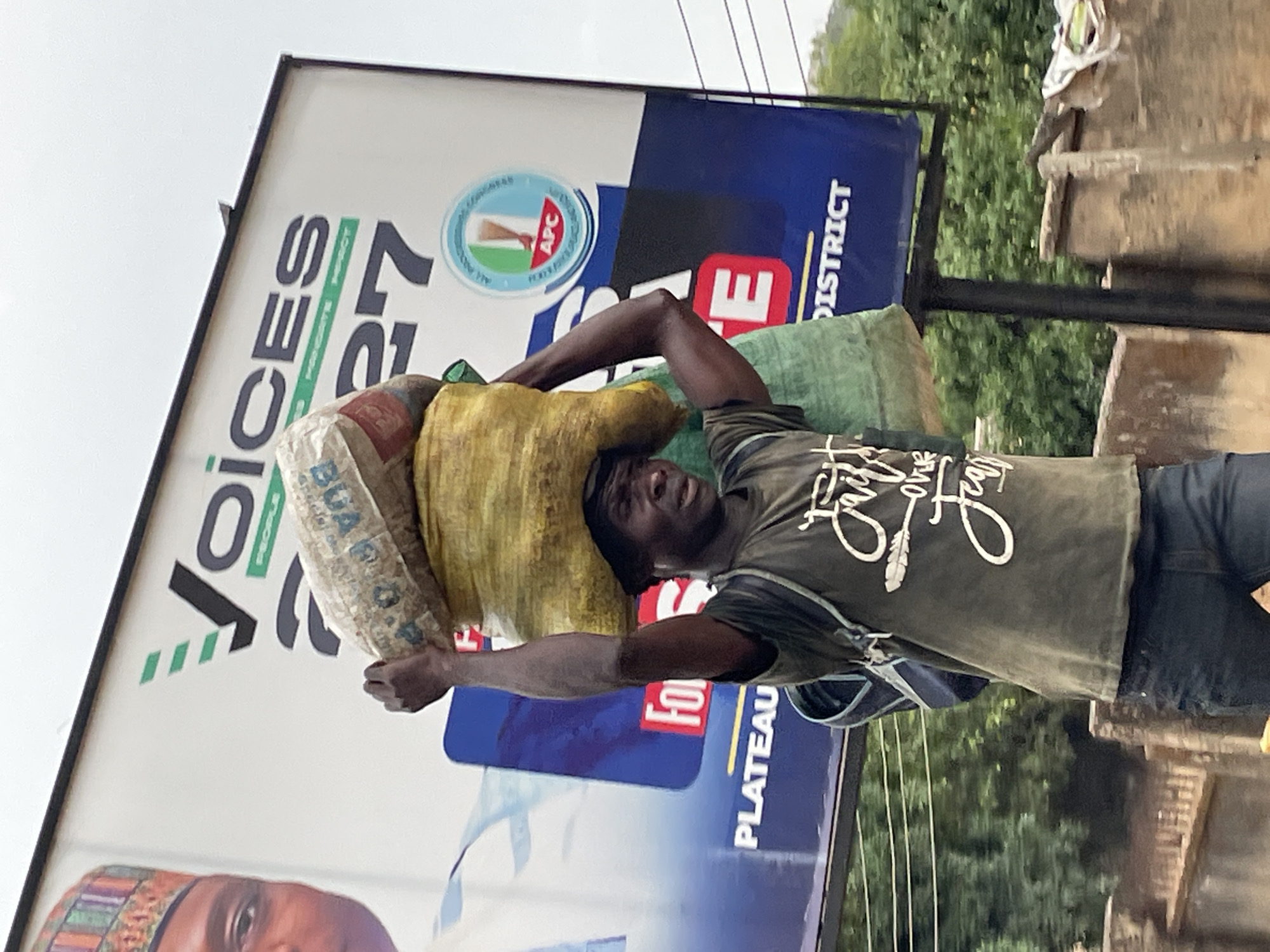 The image, captured in Jos, Nigeria, shows a man carrying heavy loads on his head, set against a backdrop dominated by a political billboard. The man is positioned centrally, facing slightly towards the right, with multiple sacks balanced expertly on his head. One visible sack is white with the partial text "BUA CEMENT," while another is yellow/tan, and a green one is visible underneath. He wears a dark green t-shirt with white script that appears to read "Faith over Fear," dark trousers, and a dark cap or head covering. His facial expression conveys a sense of effort or concentration as he transports his load, characteristic of daily labor.
Behind the man, a large, vertically oriented billboard displays political campaign material. Prominent text on the billboard includes "VOICES" and "2027." The logo of the "APC" (All Progressives Congress), a major Nigerian political party, is clearly visible, along with the words "FOR PLATEAU DISTRICT," firmly establishing the local political context. A partial image of a political figure's face is visible at the bottom left of the billboard, alongside other text like "FOR" and a large red "E."
The scene is outdoors during the daytime under an overcast sky, indicated by the soft lighting and lack of harsh shadows. Lush green foliage is visible behind the billboard and the man, suggesting a natural element in the urban or semi-urban environment. Utility wires are also visible in the upper right portion of the frame. The image captures a stark contrast between large-scale political messaging and the everyday struggle and resilience of an individual engaged in physical labor.