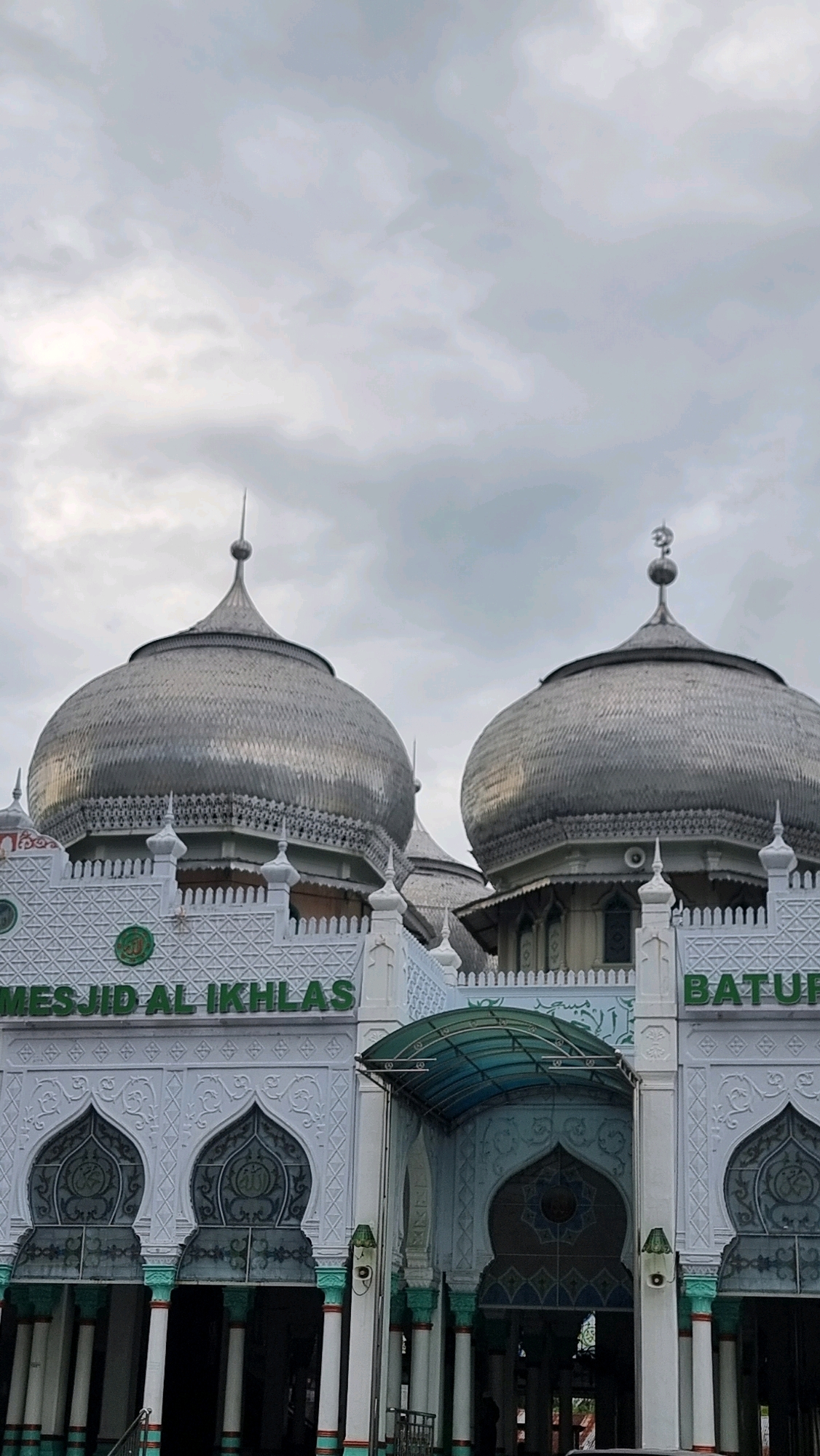The image shows the facade of the Masjid Al Ikhlas, located in the city of Lhokseumawe, Indonesia. The mosque features two prominent silver domes, the larger one in the foreground and a slightly smaller one to the right. The domes are intricately detailed with a scaled pattern and ornate trim below. The white building facade is adorned with decorative patterns and arches, with the name "MESJID AL IKHLAS" prominently displayed in green lettering above the entrance. To the right, partially visible, is the word "Batu...". A covered walkway with a teal roof extends from the entrance. The sky is overcast with gray clouds. There are no people visible in the image.