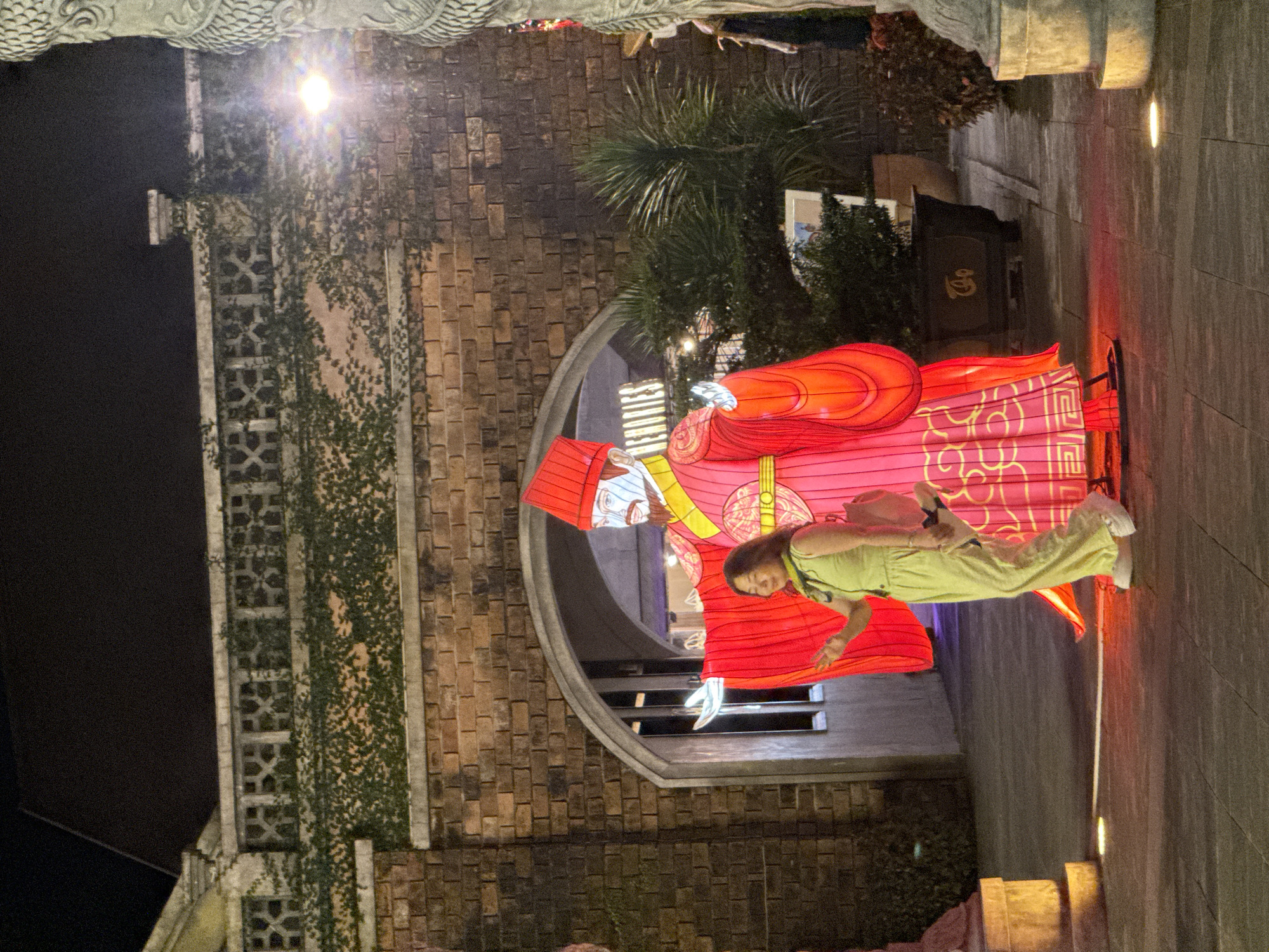 The image depicts a young woman in a lime green outfit posing next to a large, red, illuminated Chinese deity figure in Hội An Ward, Vietnam. The deity statue is positioned within a brick arched gateway. A brick building covered in climbing plants is visible on the left. The ground is paved. Artificial light sources provide illumination.