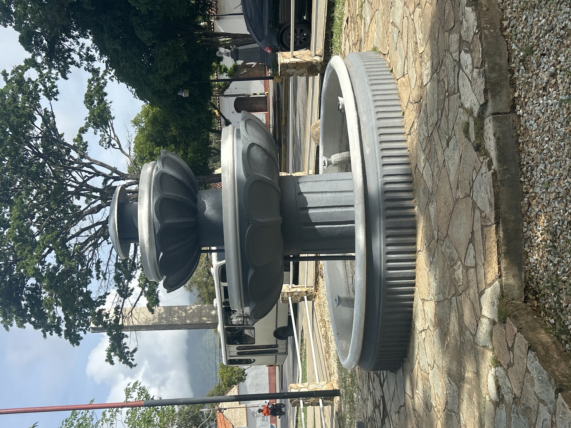 The image depicts a multi-tiered grey concrete fountain situated on a paved area composed of irregular flagstones. The fountain has a central column and several decorative tiers, with the lowest basin containing a small amount of water and a light-colored object, possibly a bird. Surrounding the fountain is a low stone wall with white railings. In the background, a white bus is visible driving on a road, with a person on a motorcycle to the left of the bus. Further back, there are buildings with red tile roofs and white walls, and a tall, slender, light-colored monument or tower. Lush green trees with a dense canopy dominate the upper portion of the image, with a cloudy sky visible through the branches. The environment suggests an outdoor public space within a town or city. This scene is located in the Plaza Bolívar of Santa Ana, Venezuela.