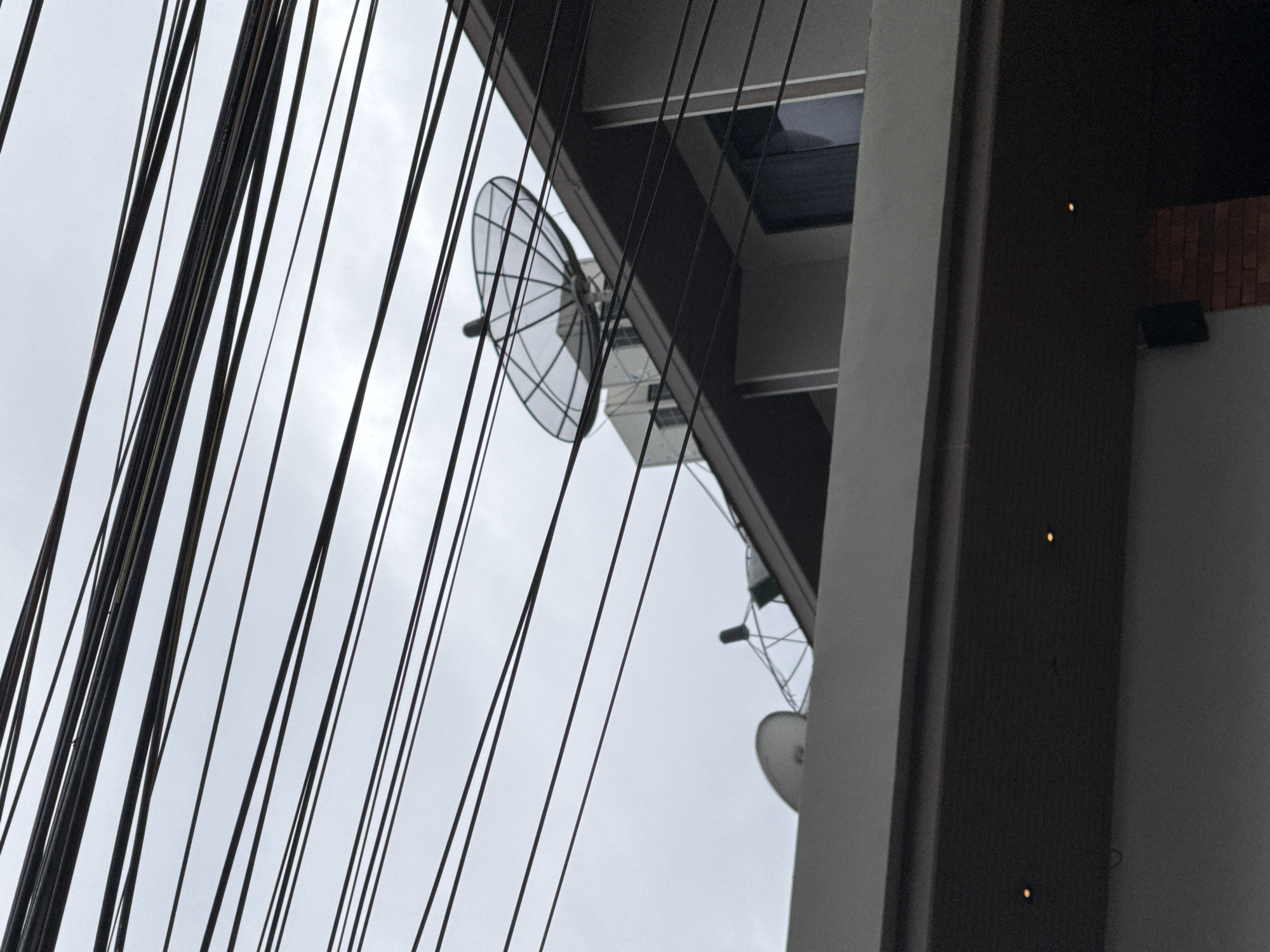 The media file displays a low-angle shot of a building's exterior, partially obscured by a dense network of dark electrical wires running diagonally across the frame. The building appears to be modern, with clean lines and a neutral color palette. A satellite dish is visible mounted on the side of the building, suggesting a location where such technology is common for communication or entertainment. In the background, the sky is overcast and a pale gray, indicating a cloudy day. There are also some small, yellowish lights visible on the dark facade of the building, possibly exterior lighting. The scene is likely set in an urban or semi-urban environment, given the presence of the wires and the multi-story building. The overall mood is somewhat stark due to the dominance of the wires and the muted colors. There are no people or specific activities depicted, making it a still-life composition focusing on urban infrastructure. The location context of Karon, Thailand, suggests a tropical or subtropical climate, but the weather in this particular shot appears to be cloudy.