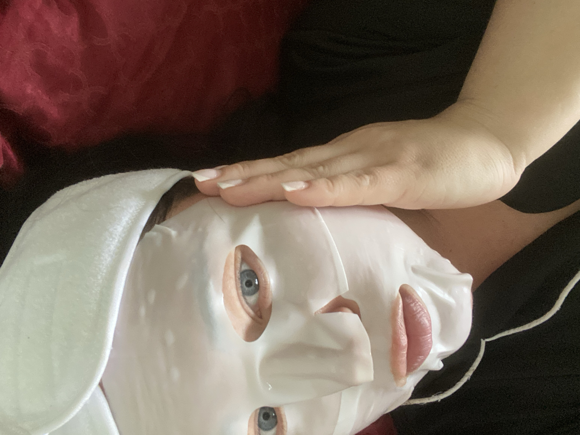 A woman is lying down, wearing a white sheet face mask and a white headband. Her hand is resting on her forehead. She has blue eyes and painted fingernails. The background includes a dark garment and a maroon pillow. The location is unknown, and there is no information available to suggest that this picture was taken in Sochi, Russia.