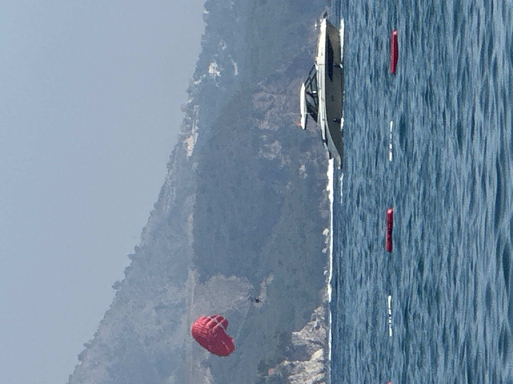 A parasailer, suspended from a red parachute, is seen in mid-air against a hazy mountain backdrop and a calm blue sea. The parachute is taut and angled, indicating the parasailer is actively being towed. Below, a sleek, light-colored motor yacht cruises across the water, leaving a white wake. In the foreground, several buoys are visible, marking an area of the sea. The mountainous terrain behind the parasailer is covered in lush vegetation, and the sky is a pale, hazy blue. The scene appears to be a recreational area, likely a coastal resort, possibly in the vicinity of Boreti, Montenegro, given the characteristic Mediterranean landscape and popular water sports activities.