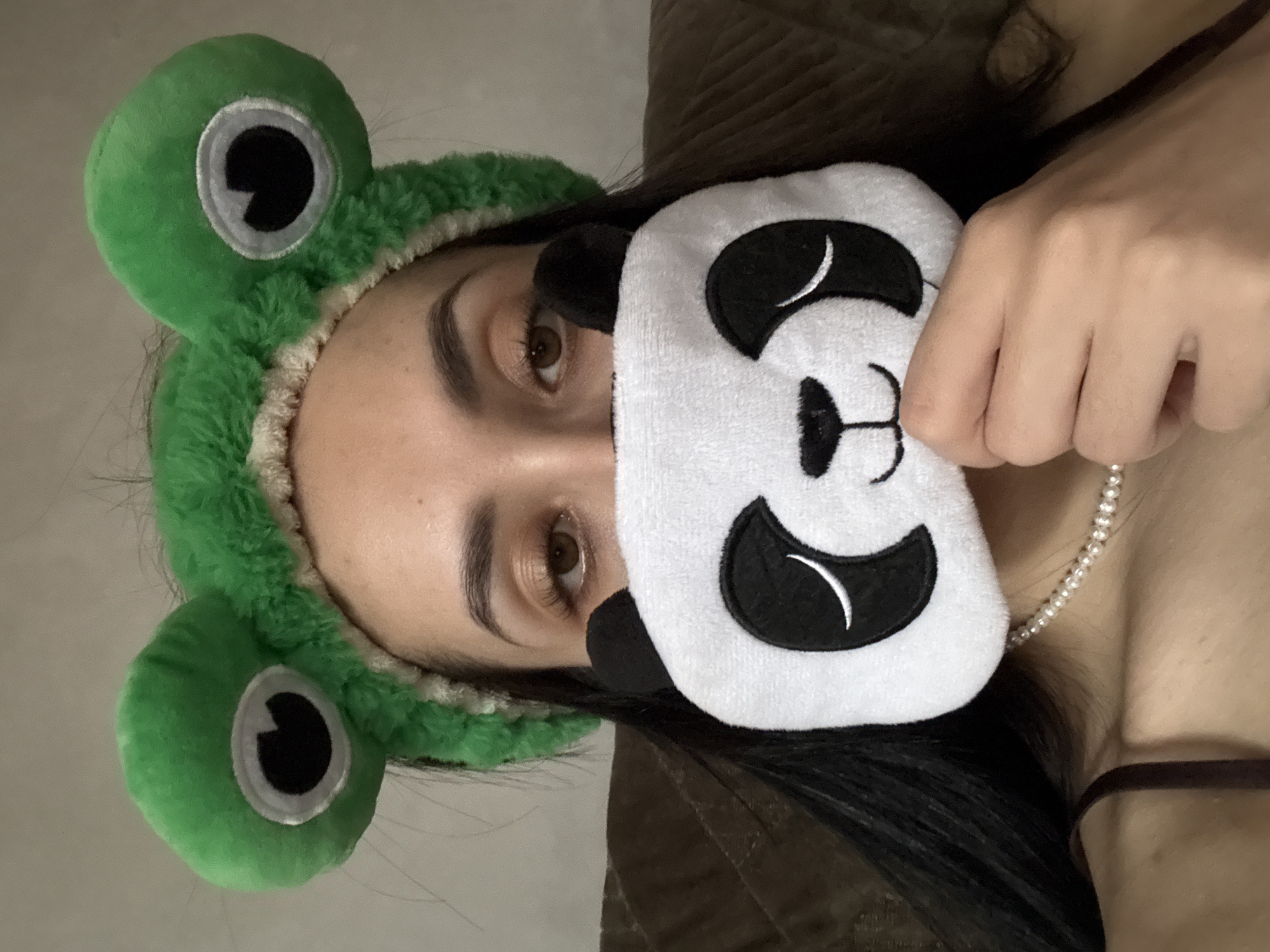 An indoor, horizontally oriented close-up shot features a person, likely female, with dark hair and brown eyes. The person wears a bright green, plush headband designed to resemble a frog, complete with large, prominent eyes on top. She holds a white and black plush eye mask, shaped like a panda's face, partially covering the lower half of her own face. One of her hands is visible gripping the eye mask. A pearl necklace is worn around her neck, and a thin clothing strap can be seen near her shoulder. The background is softly lit and includes a lighter, plain area, possibly a wall, and a darker, textured section, suggesting bedding or furniture. The visible eye shows a neutral and relaxed expression. The scene suggests a casual, playful, and comfortable moment of leisure or self-care. No text is visible in the image. The context for this image is Votkinsk, Russia.