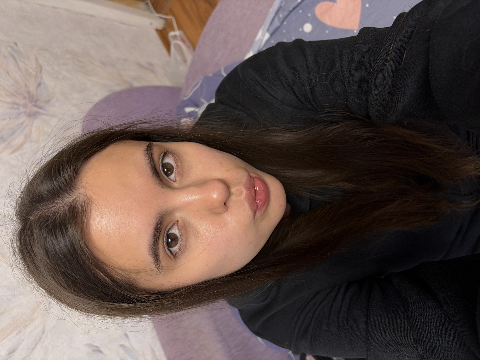 A selfie-style photograph shows a young woman with brown hair and eyes. She is wearing a black top. The background includes a bed with lavender and patterned sheets. It is impossible to determine the location of the city of Chaikovsky, Russia. The image provides no contextual clues to ascertain that location.