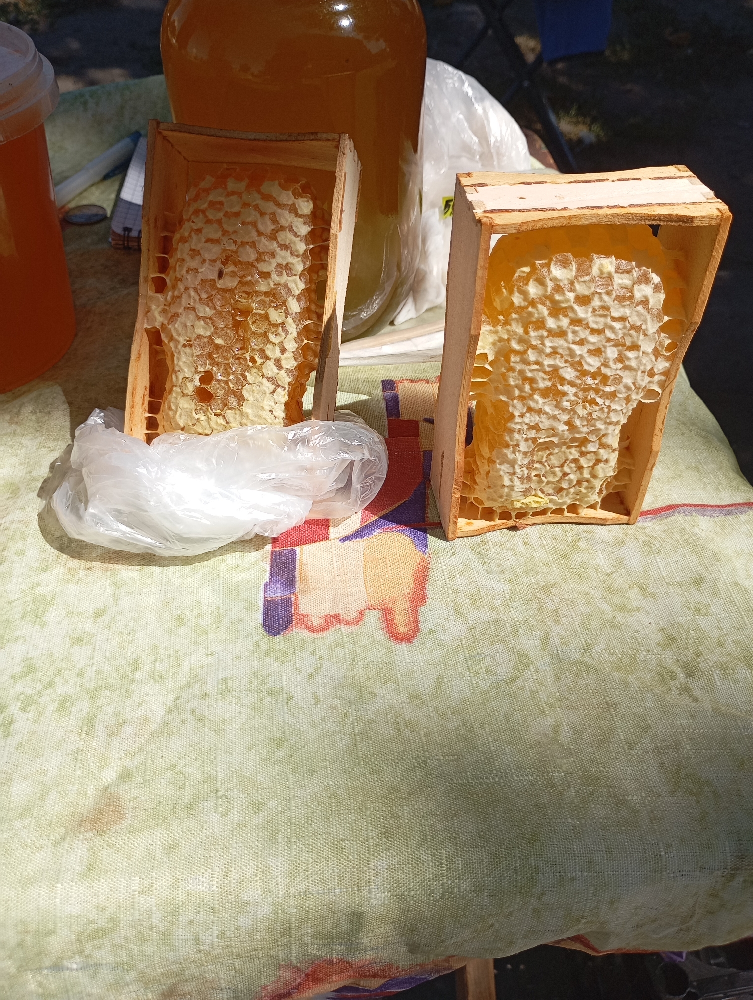 Two wooden frames, each containing a piece of honeycomb filled with honey, are displayed on a patterned tablecloth. The frames are positioned in the foreground, with the honeycomb in the left frame appearing slightly more filled and with some darker cells. Behind these frames, a large glass jar filled with honey is visible. To the left of the frames, a translucent plastic bag lies on the tablecloth. The scene is set outdoors, with sunlight casting shadows. The tablecloth has a light green background with a repeating pattern of abstract shapes in red, purple, and yellow. The backdrop includes blurred outdoor elements, suggesting a market or roadside stall setting. This image was captured in Horishni Plavni, Ukraine.