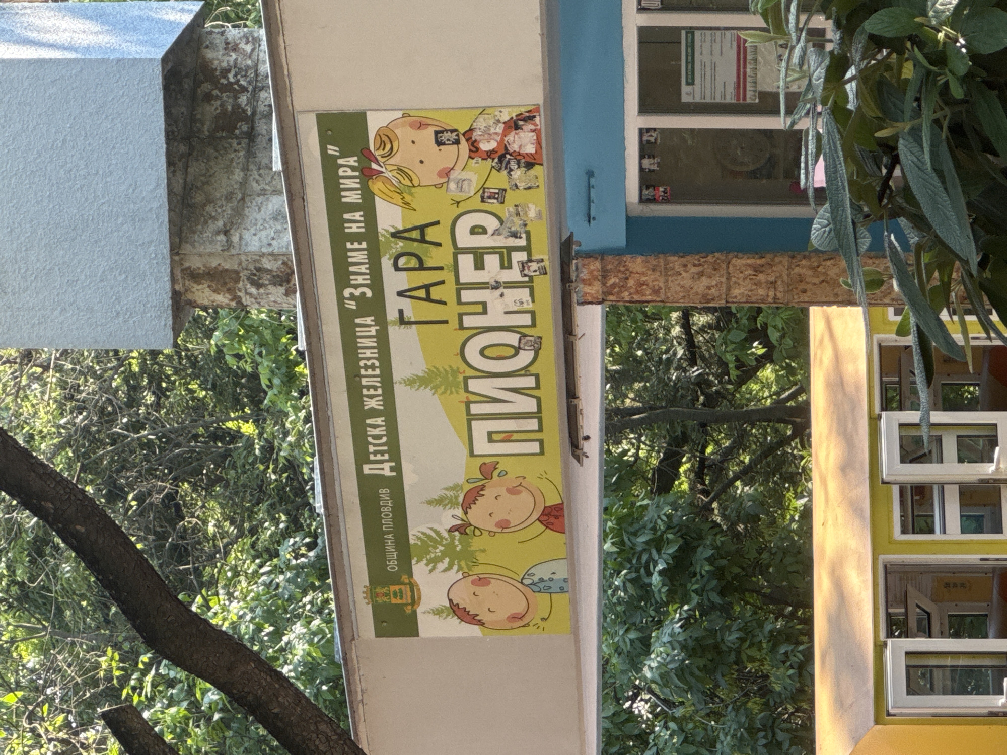 The image shows a sign for "Gara Pioneer" in Plovdiv, Bulgaria. The sign is attached to a building. It has cartoon illustrations of children on a green and yellow background. The building is partially visible and has windows. Foliage and trees are visible in the background.