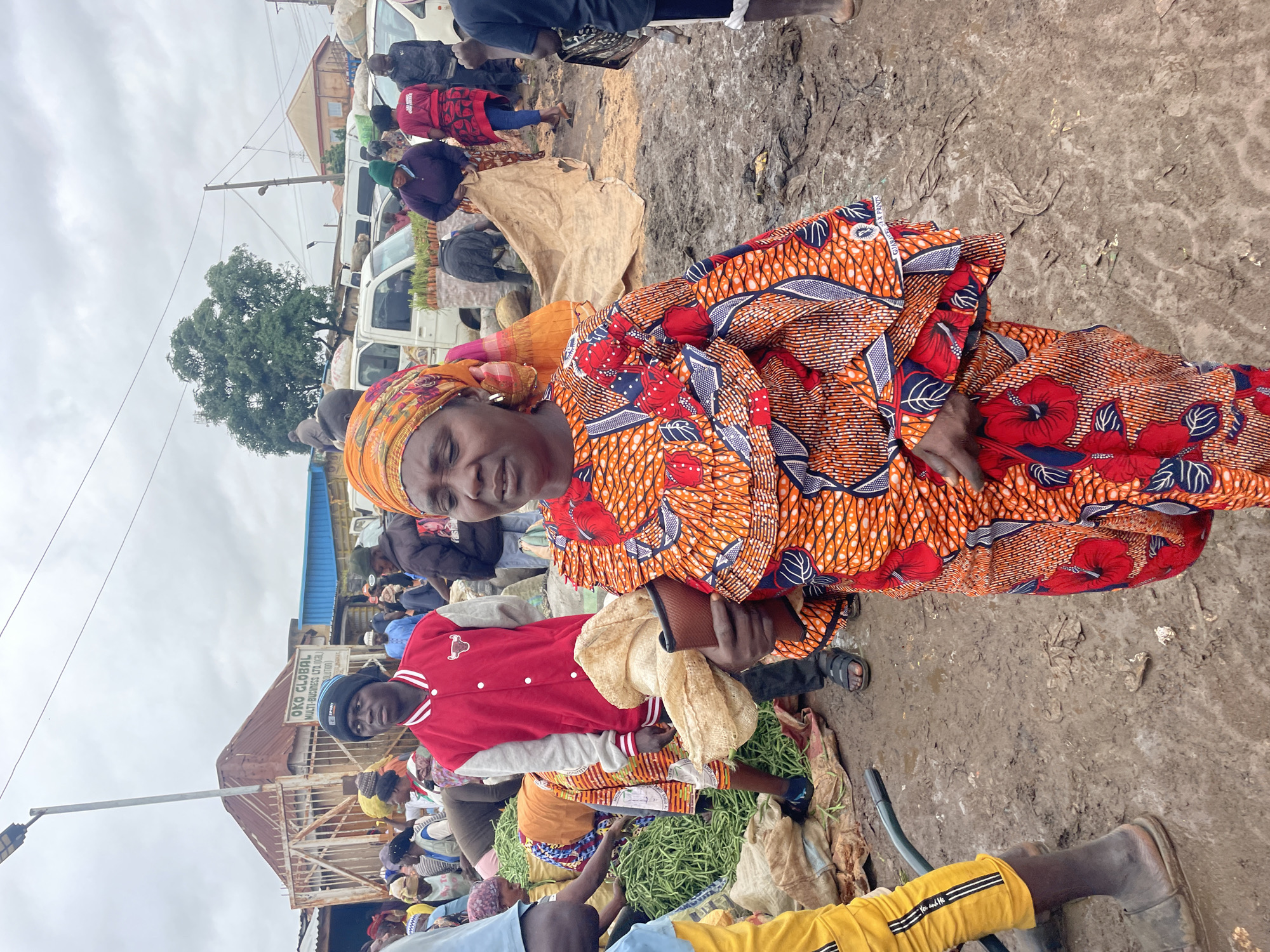 The image depicts a bustling outdoor market scene in Gura Top, Nigeria. In the foreground, a woman wearing a vibrant orange and red patterned dress and headscarf is the central subject. She is holding a brown wallet and a piece of light-colored fabric. Behind her, a young man in a red and grey bomber jacket and a black beanie stands looking to his left. The market is crowded with people, some of whom are interacting with piles of green vegetables. Various vehicles, including vans and a truck, are visible in the background. The ground is muddy, indicating recent rain or a generally damp environment. Structures, including a building with "OKO GLOBAL MULTI-BUSINESS LTD." signage, are also present. The sky is overcast, suggesting cloudy weather.
