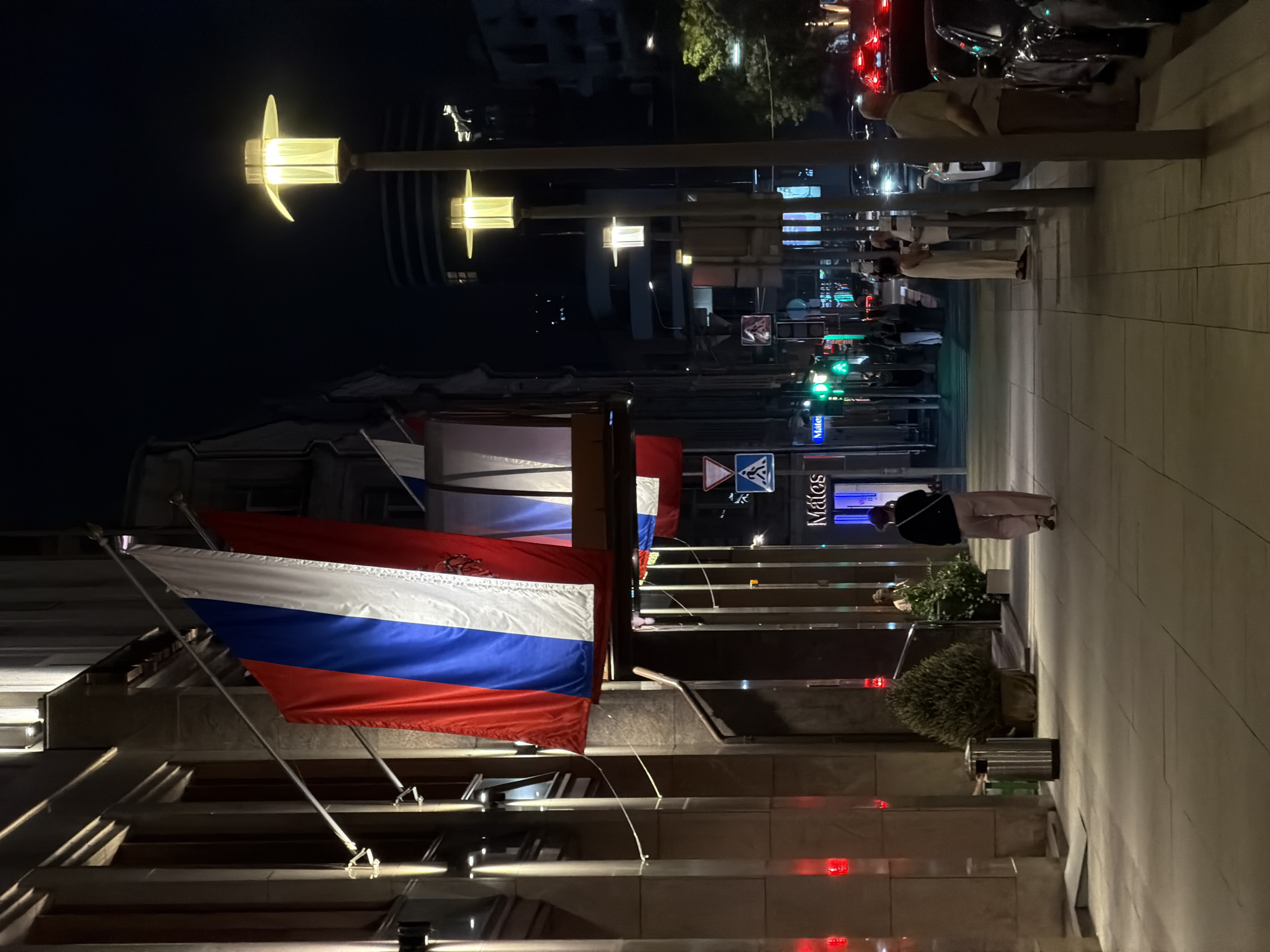 The image shows a night scene in Moscow, Russia. The Russian flag is prominently displayed on a building. Pedestrians walk along a sidewalk lined with streetlights. Cars are visible in the background, and traffic signals are lit. A sign reading "Mates" is also visible.
