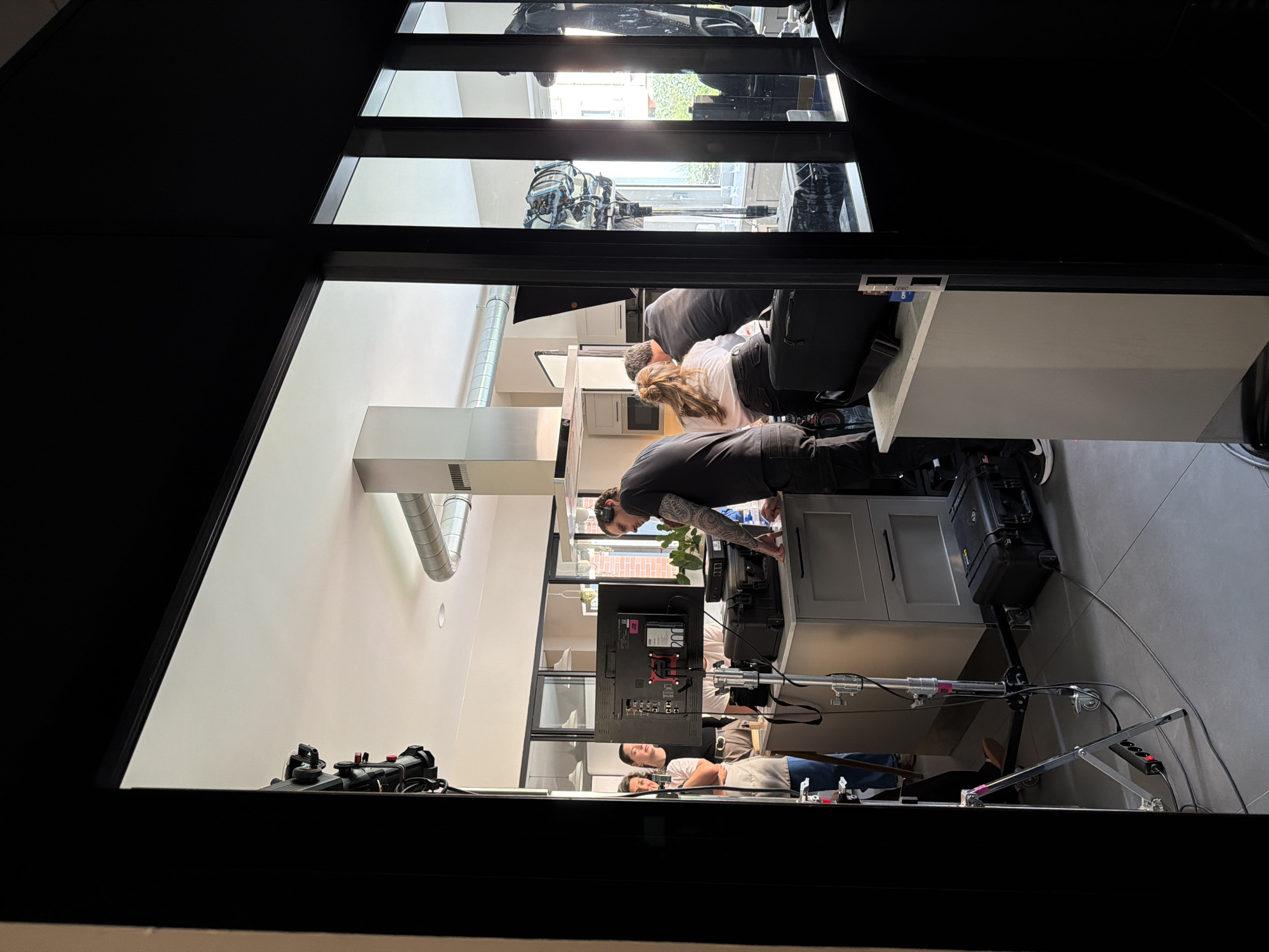 The image shows an interior scene, likely in a residential or commercial space, as viewed through a dark doorway or aperture.
Setting and Environment: The room inside has a modern aesthetic with white walls and gray flooring. A kitchen area is visible, featuring white cabinets, drawers, and a stainless steel ventilation system. A television screen with production equipment, cords, and camera equipment is set up in the kitchen. Daylight enters from windows in the distance.
People: Several people are present in the room, appearing to be involved in a film or video production. One individual with a tattoo is seen bending over, interacting with equipment on a counter. Two other individuals are lying on the floor. Another pair of people are in a kitchen setup behind the counter, and another obscured.
Objects and Equipment: Camera equipment, including tripods, cables, monitors, and a black equipment case, are present.
Actions and Interactions: The individuals appear to be setting up or operating film or video production equipment.
Location: While the request specifies Aubervilliers, France, this information is not visually apparent from the image alone.