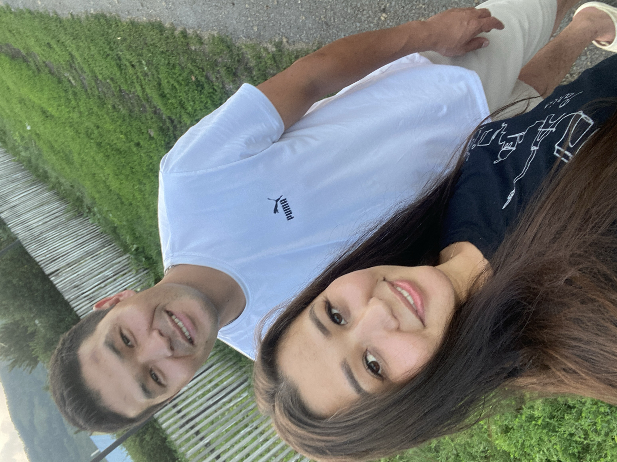 A waist-up, outdoor selfie features a man and a woman in Maloarslangulovo, Russia. The man, positioned behind the woman, smiles directly at the camera, his face in sharp focus. He wears a white t-shirt with a Puma logo on the left chest and tan shorts. The woman, closer to the camera and slightly to the left, looks slightly away from the lens with a gentle smile. She has long, dark brown hair and wears a black t-shirt with a white graphic design. They are standing on a gravel path next to a grassy area bordered by a wooden fence. In the background, a treeline and a soft, overcast sky can be seen. The lighting suggests it is late afternoon or early evening.