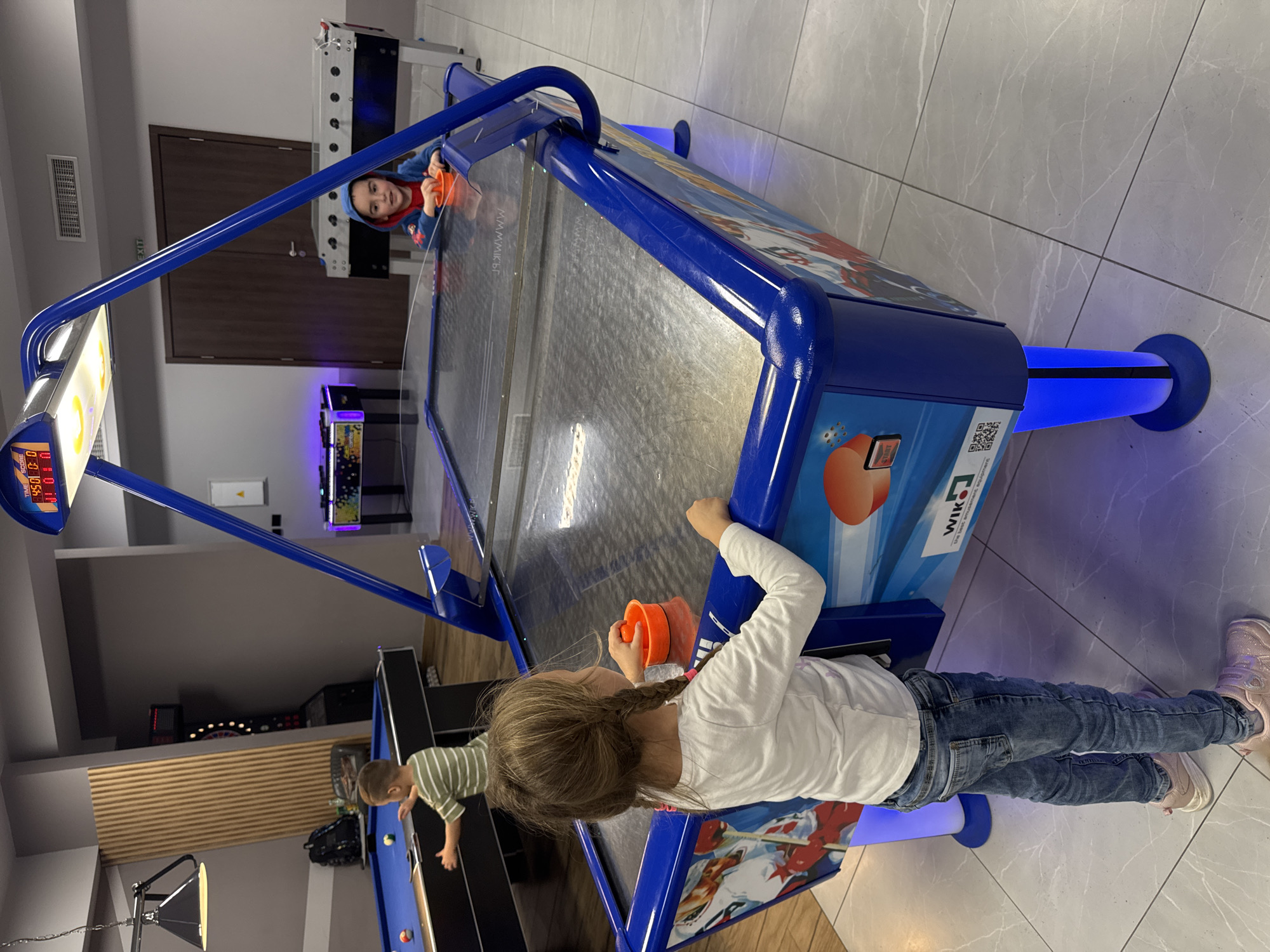 The image shows a game room in Kranevo, Bulgaria. Two children are playing on an air hockey table. One child, a girl, is standing on the near side of the table, holding an orange puck and looking into the game. She is wearing a white long-sleeved shirt, blue jeans, and pink sneakers. Her hair is blonde and braided. The other child, a boy, is on the far side of the table, also holding an orange puck. He is wearing a blue hooded sweatshirt. In the background, another child, a boy wearing a green and white striped shirt, is playing on a blue pool table. There is a foosball table in the background as well. The floor is tiled in gray.