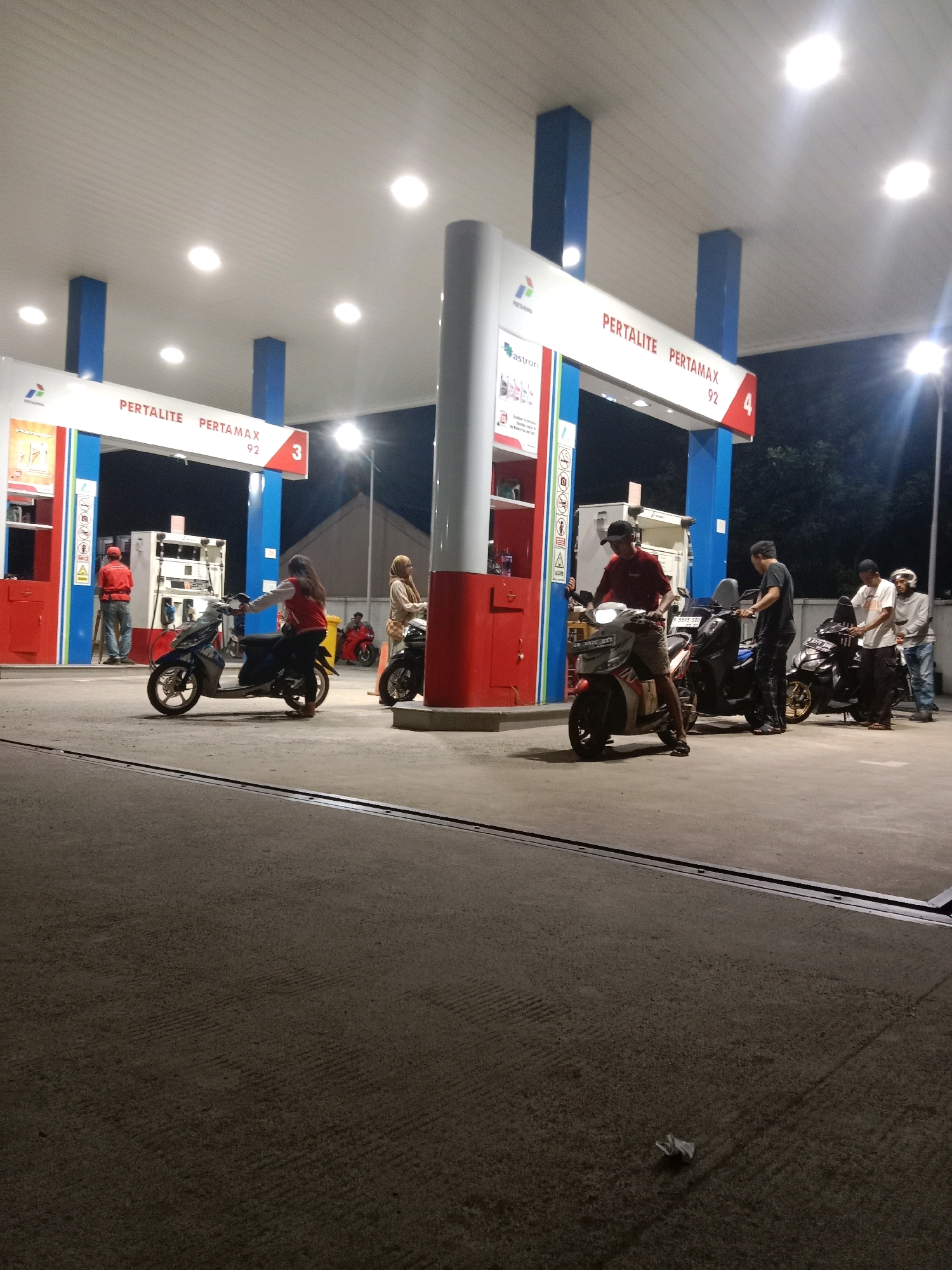 The image captures a nighttime scene at a Pertamina gas station, likely located in South Tangerang, Indonesia. The station is illuminated by overhead lights, casting a bright glow on the covered forecourt. A white awning, supported by blue pillars, extends over the fueling area. Signs above indicate "PERTALITE" and "PERTAMAX 92".
Several individuals are present. On the left, a person wearing a red jacket and a cap stands near a fuel pump. To the right of the pump, a woman on a scooter, also wearing a red jacket, is stopped. Further to the right, a man in a maroon shirt is on his scooter, seemingly preparing to refuel or having just refueled. Behind him, another man in a dark shirt stands next to a black motorcycle. Two more individuals are visible in the background, one in a white shirt and another in a light-colored jacket, both standing near motorcycles. A woman wearing a hijab is also present, standing next to a black motorcycle.
The ground surface appears to be asphalt or concrete. In the foreground, a crumpled piece of paper is visible on the ground. The background shows a dark, possibly residential, area with building structures visible. The overall atmosphere suggests a typical evening at a busy gas station.