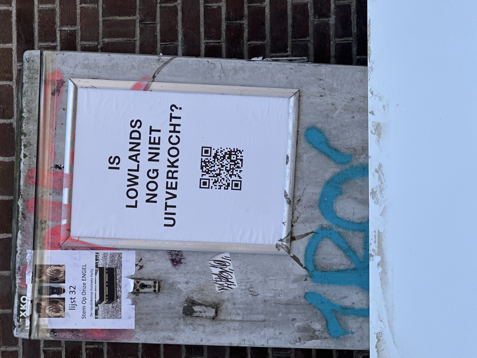 Here is a detailed description of the image:
The image shows a close-up of a grey metal box or cabinet attached to a brick wall. The metal surface is weathered with traces of graffiti. A framed white sign is affixed to the metal box. The sign has the question "IS LOWLANDS NOG NIET UITVERKOCHT?" printed in bold, black letters. Below the question is a QR code.
Under the sign, a ripped and weathered election poster labeled "lijst 32 Stem Op Onze ENGEL De republikeinse Politieke Partij" is stuck to the metal.
The location is indicated by the Dutch text on the signs, suggesting it is in the Netherlands, perhaps in Amsterdam. However, the image doesn't offer any additional clues to confirm its specific location in Amsterdam.