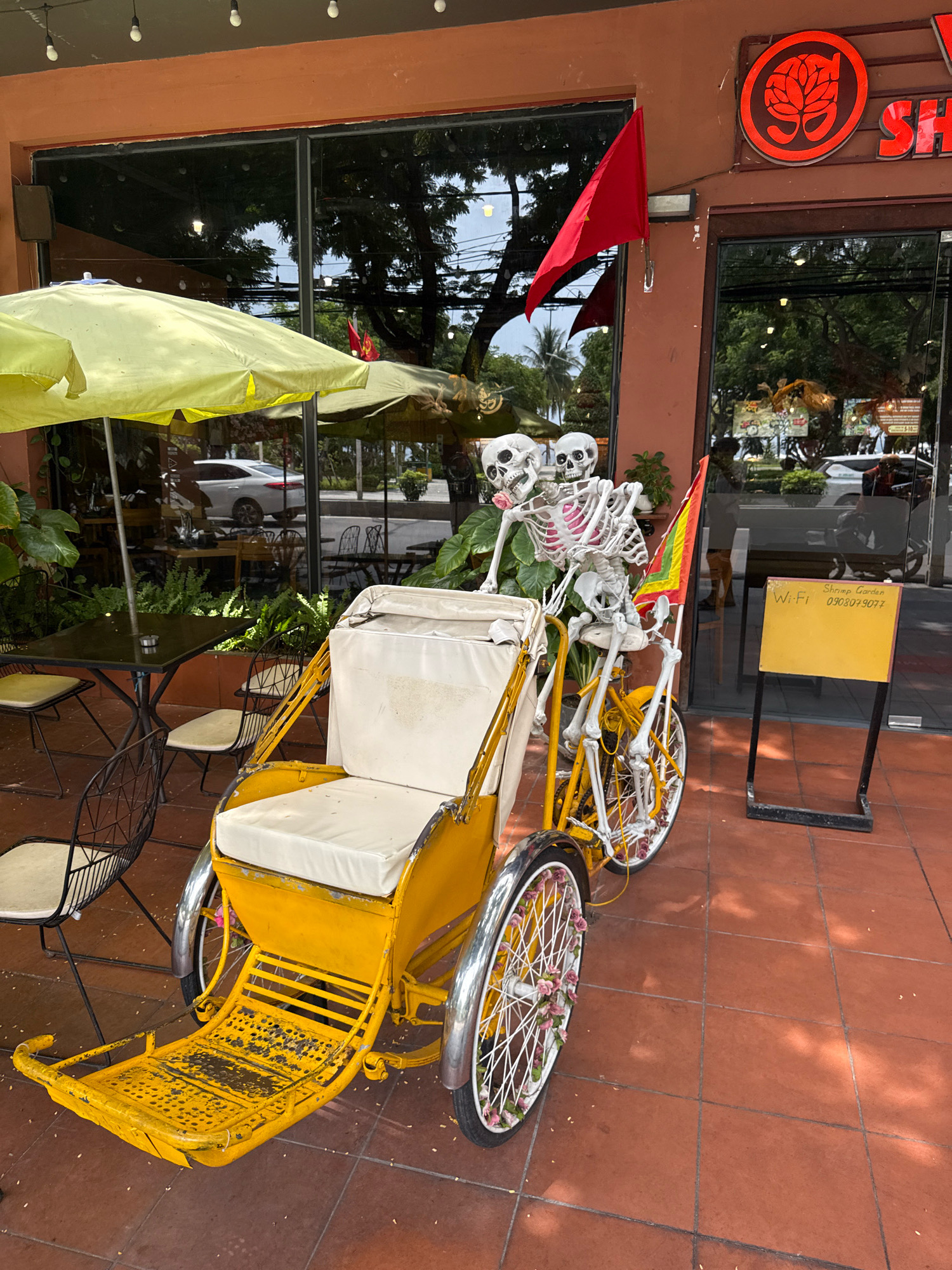 The image depicts a street scene in Nha Trang, Vietnam, likely outside a commercial establishment. In the foreground, a bright yellow, three-wheeled rickshaw is prominently displayed. Two skeleton figures, posed as if riding, are seated on the rickshaw. One skeleton appears to be holding onto the other, with one arm around its companion. A colorful flag, red and yellow, is attached to the rickshaw.
Behind the rickshaw, a large window reflects the surroundings, showing trees and possibly other vehicles. A bright yellow umbrella provides shade over an outdoor seating area with tables and chairs, suggesting a restaurant or cafe. A red flag is mounted on the building's facade. To the right, a yellow sign with black text is visible, displaying "Wi-Fi," a phone number, and "Shrimp Garden." The ground is paved with reddish-brown tiles. The overall impression is that of a decorated storefront with an unusual display.