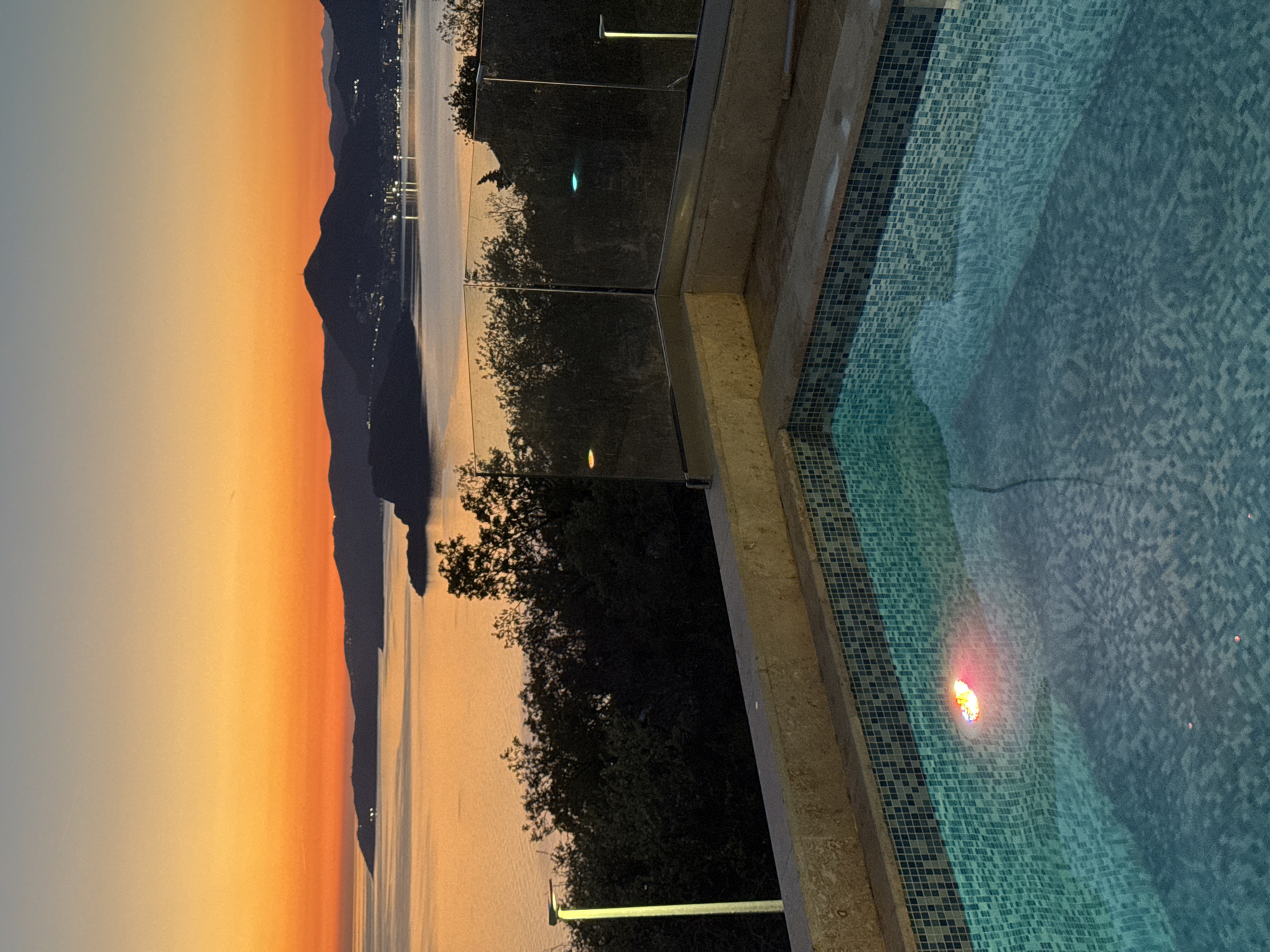 The image depicts a view from a swimming pool in Blizikuće, Montenegro, at sunset. The pool is tiled with small, light-blue mosaics, and the water reflects an underwater light emitting a reddish glow. A glass railing separates the pool from a view of the Adriatic Sea, mountains silhouetted against an orange-tinted sky, and trees. The scene is lit by the natural light of sunset.